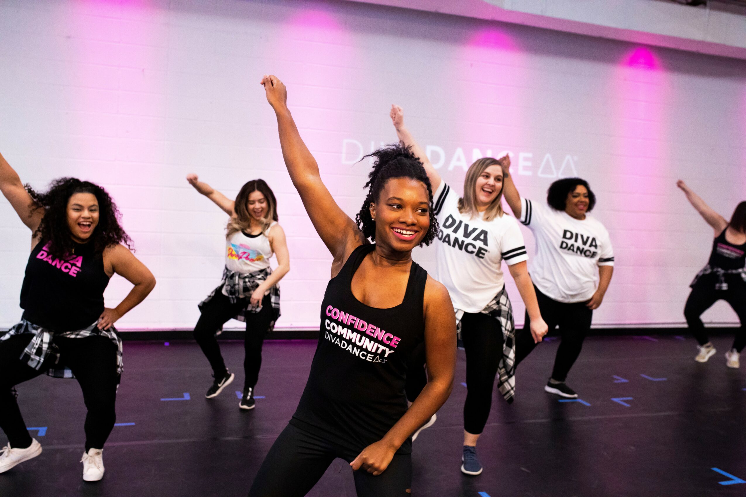 Group of women dancing on stage with pink lighting, smiling, wearing casual clothes, some with