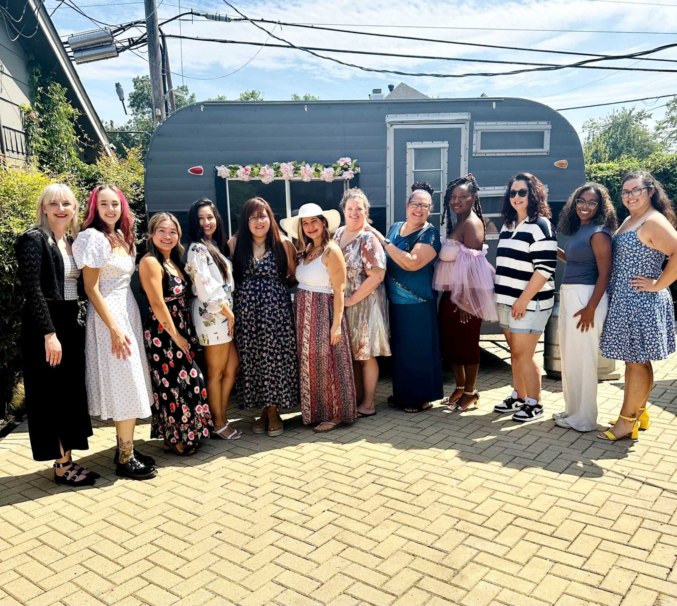 Group of women standing outdoors in front of a trailer, smiling, with trees and power lines in the background.
