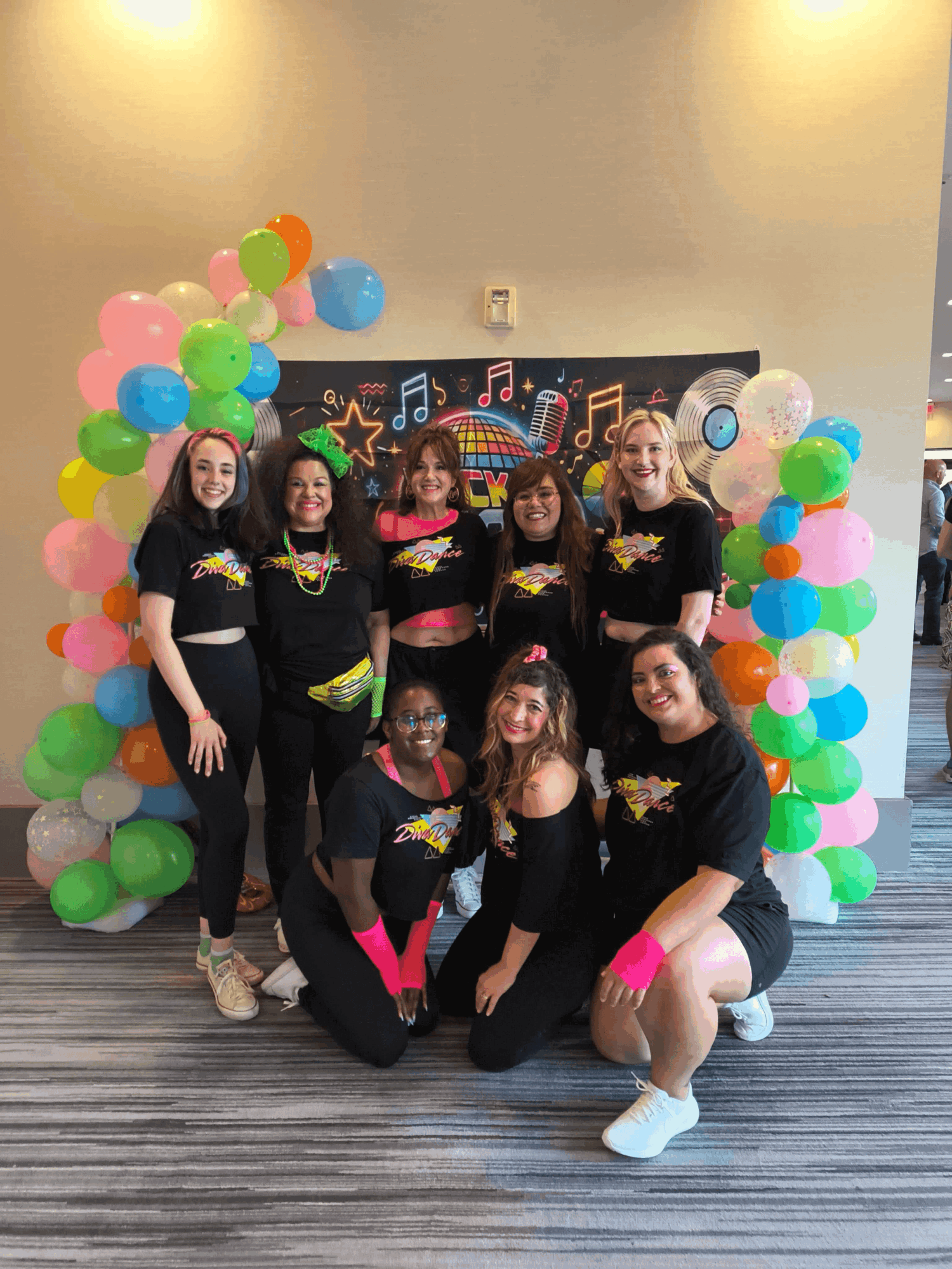 Group of seven women in black shirts with colorful designs, standing and kneeling in front of a balloon arch and a colorful backdrop.