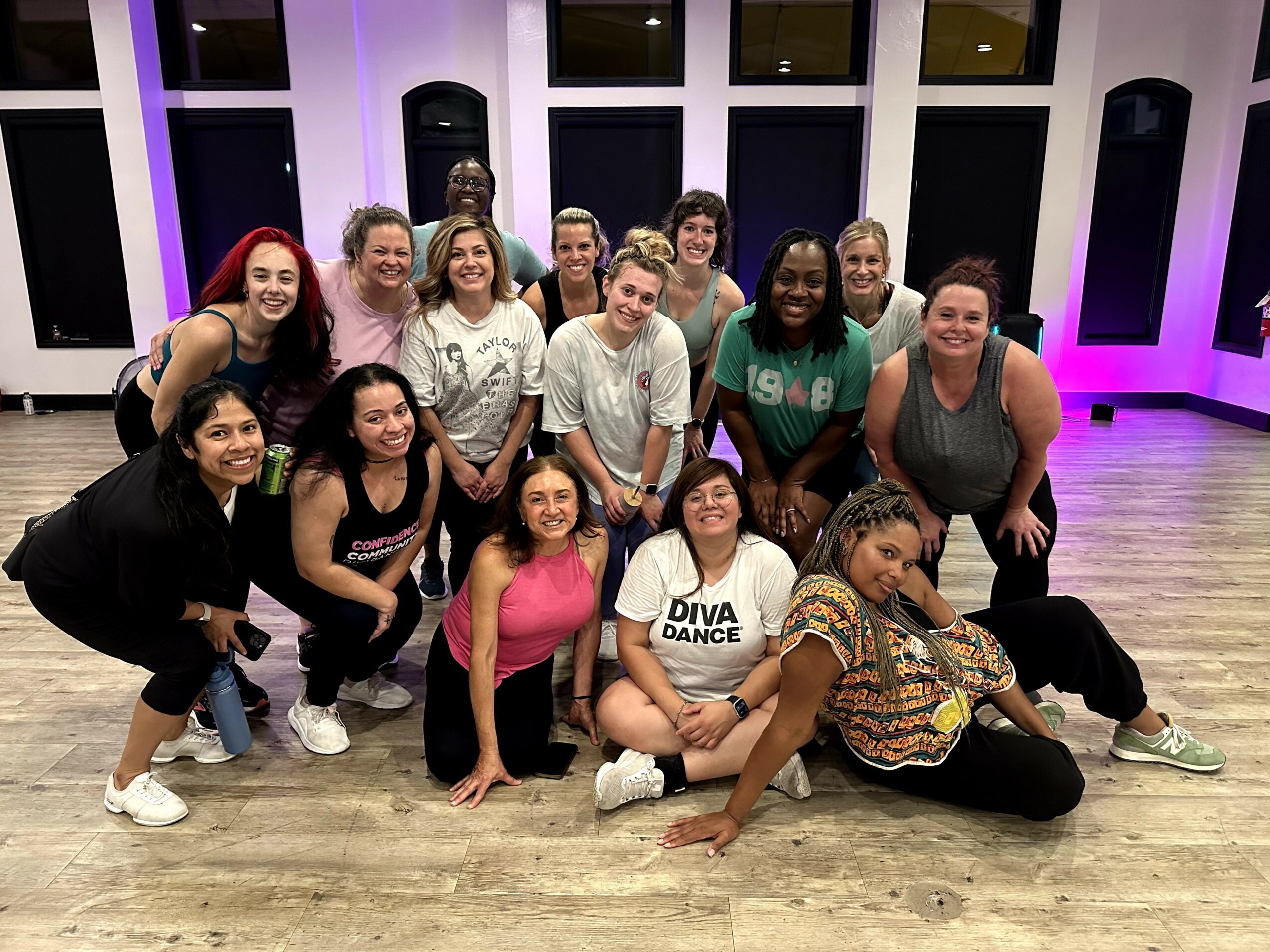 Group of 15 women posing indoors with large windows and purple lighting, some sitting and some standing, smiling at the camera.