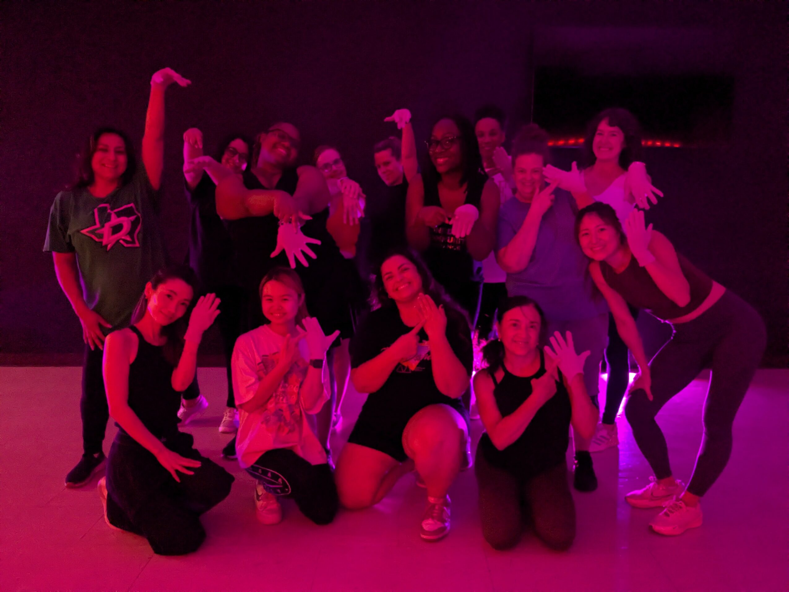 Group of people posing together in a dark room with purple lighting, some making gestures or poses, some standing or kneeling.
