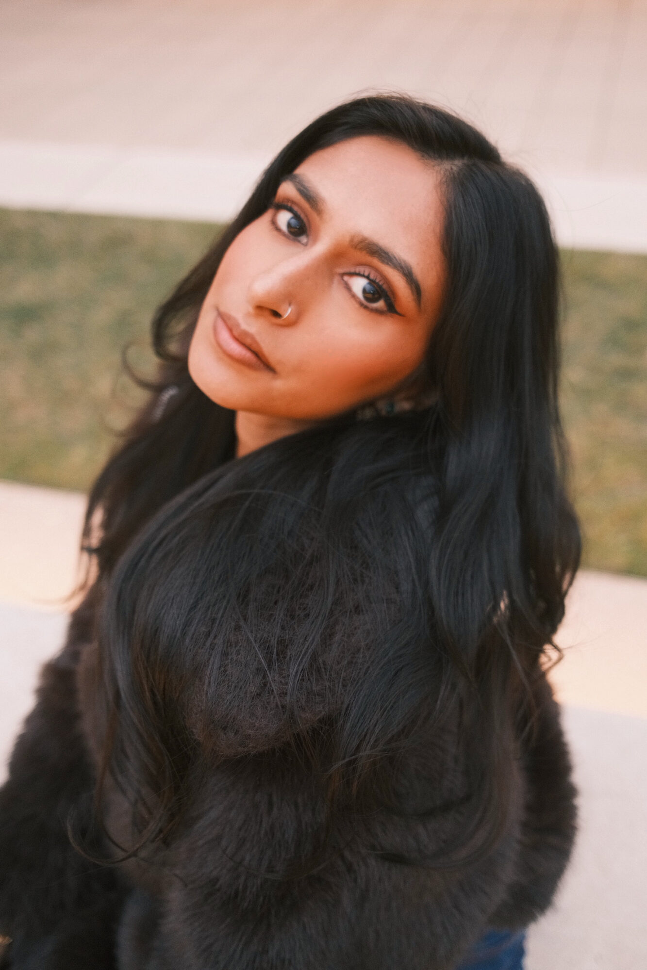 Close-up of a woman with long black hair, looking at the camera outdoors, with a blurred background.
