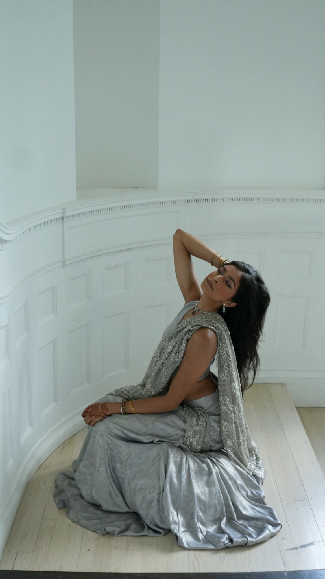 Woman in a silver dress kneeling on the floor, with one arm raised behind her head, in a white room.