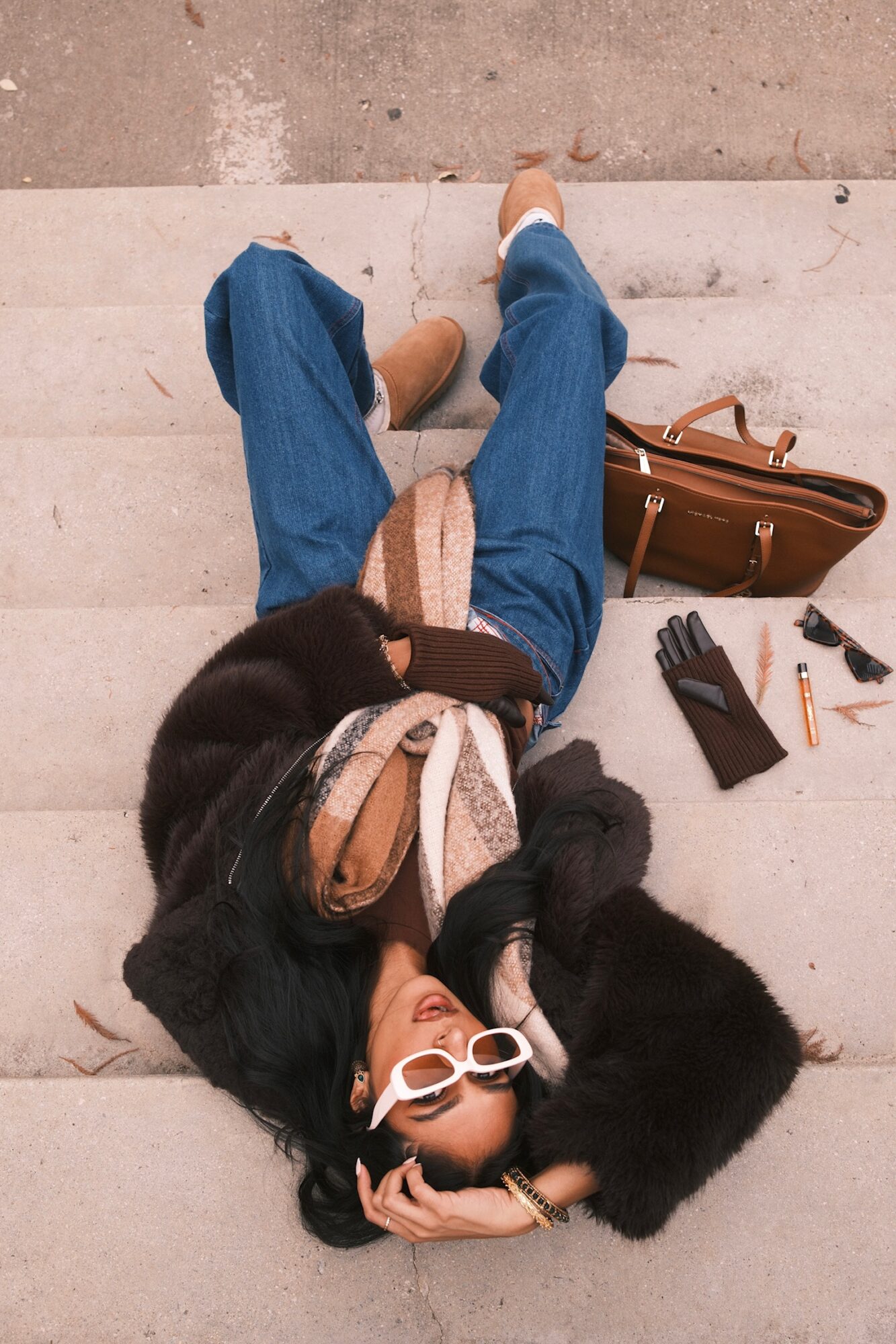 Person lying on stairs with sunglasses, black coat, jeans, and a bag nearby, holding a cigarette.