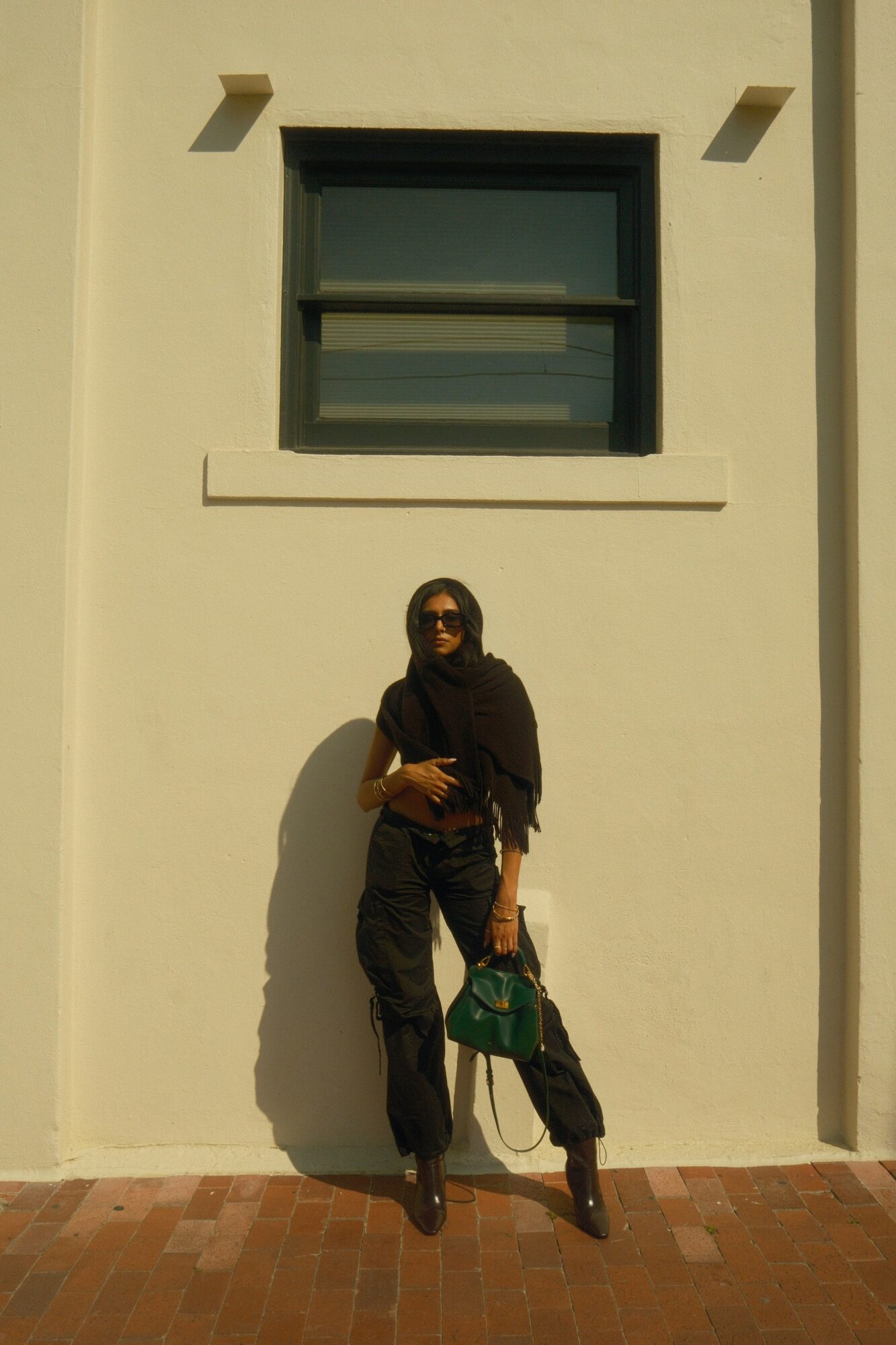 Person standing against a white wall with a window above, holding a green bag, wearing dark clothing and sunglasses.