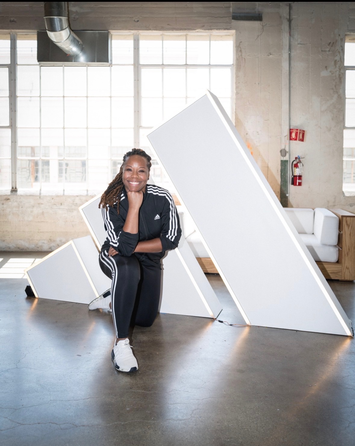 Person kneeling with arms crossed, smiling, in an industrial-style room with large windows and white geometric structures.