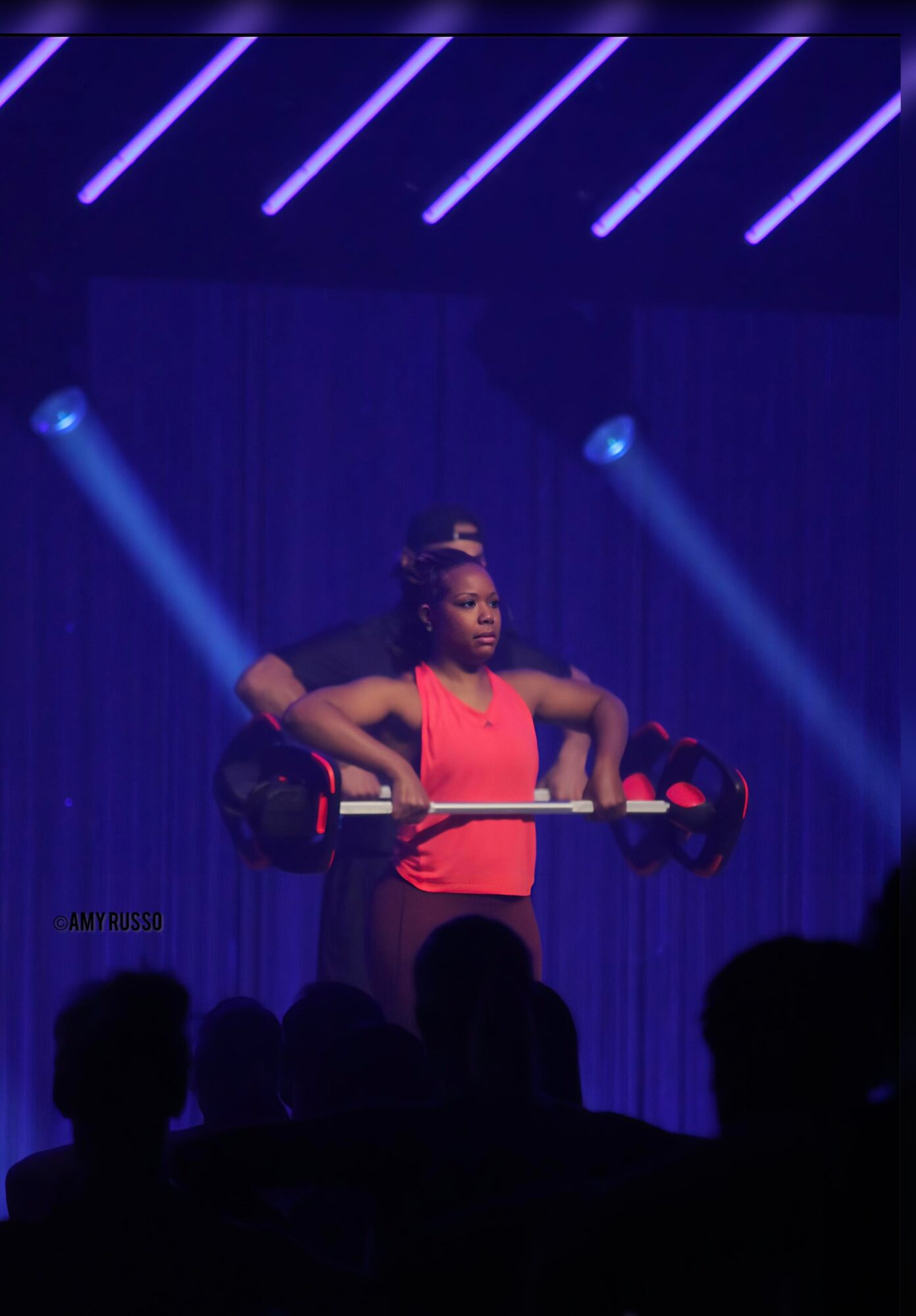 A woman lifts a barbell on stage with blue and purple lighting, audience silhouettes visible in foreground.