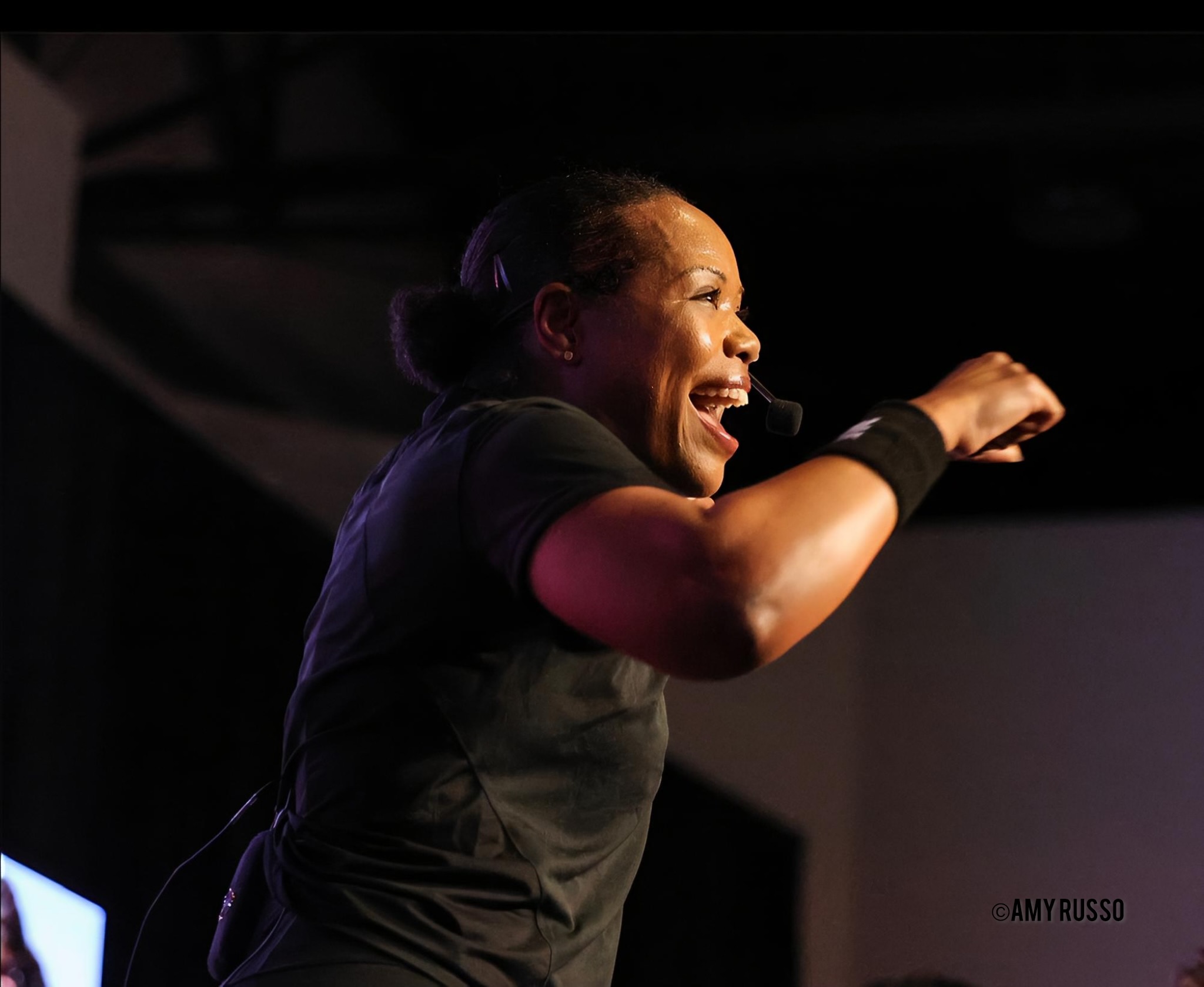 Smiling woman with hair in a bun, wearing a black shirt, raising her arm in a celebratory gesture.