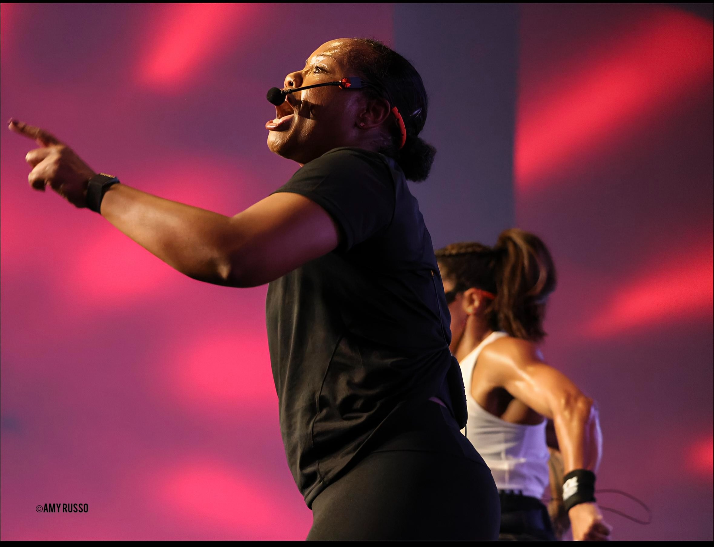 Person with glasses and ponytail pointing, on stage with pink and purple lighting, another person in background.