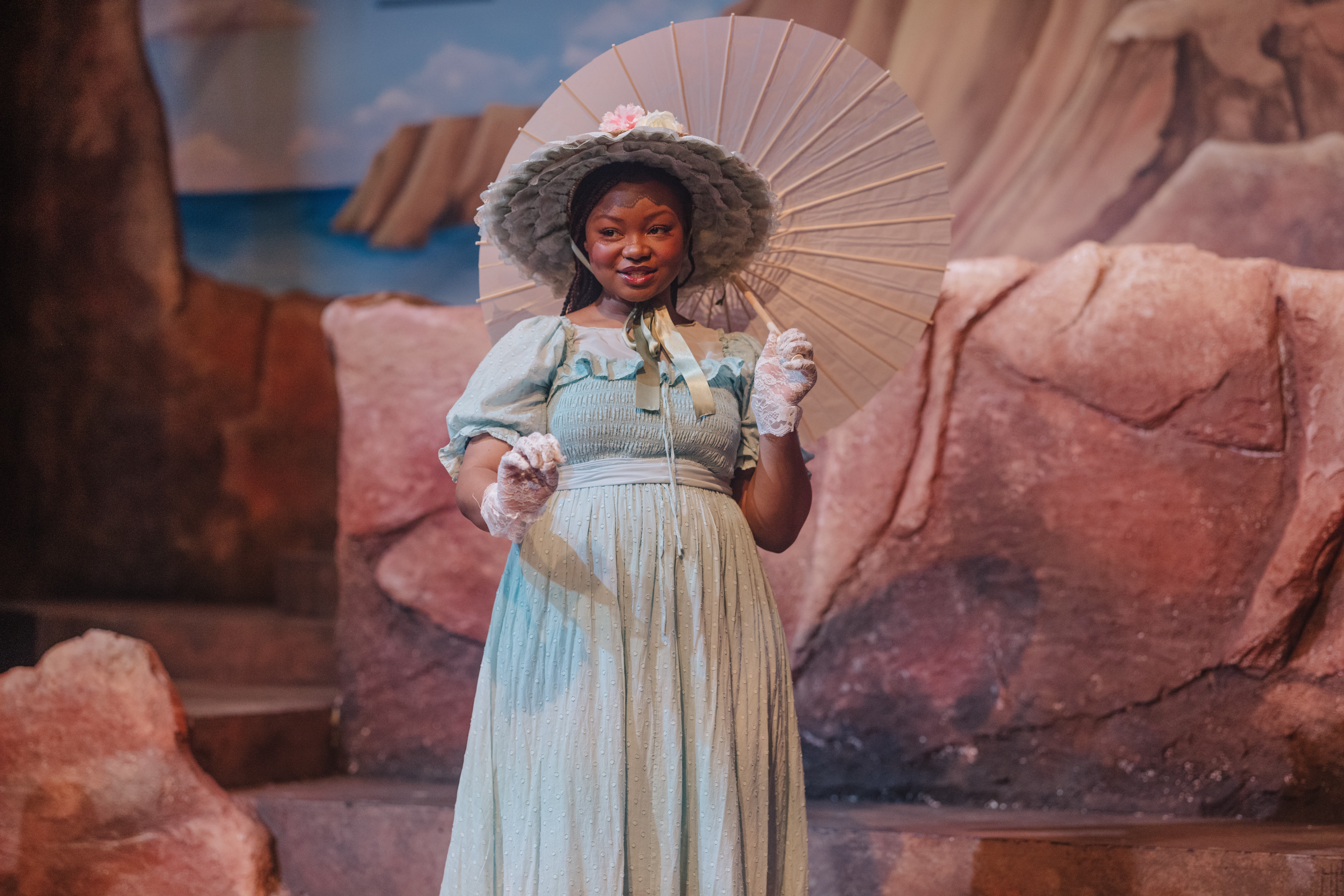 A woman in historical costume holding a parasol, standing outdoors with rocks in the background.