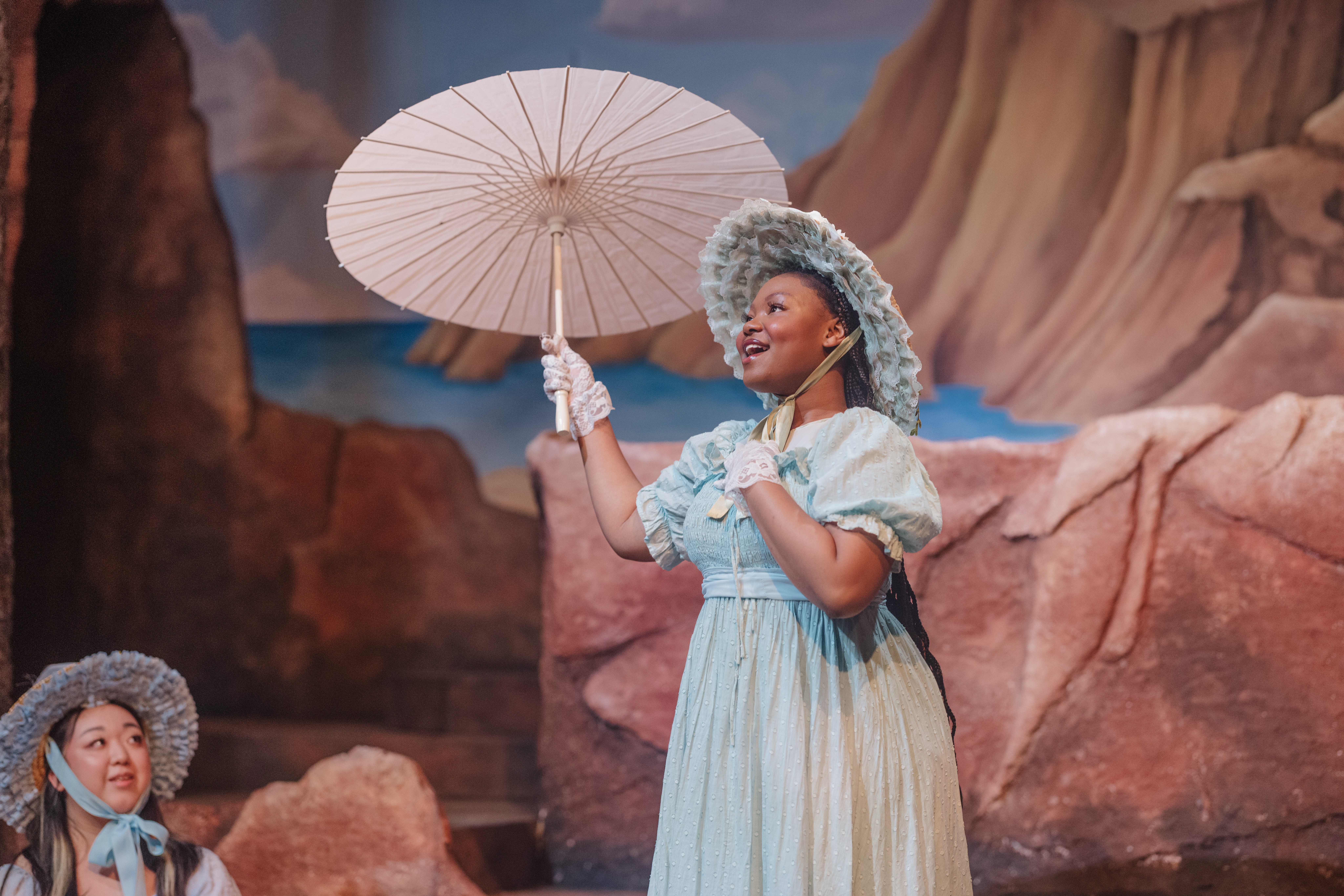 A woman in a costume with a curly gray wig holding a white parasol, standing outdoors with rocky cliffs in the background.