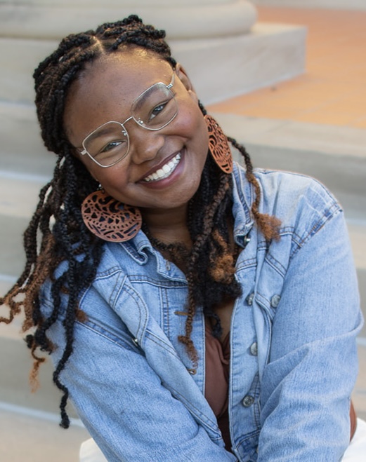 Smiling woman with glasses and earrings, wearing a denim jacket, outdoors with steps in background.