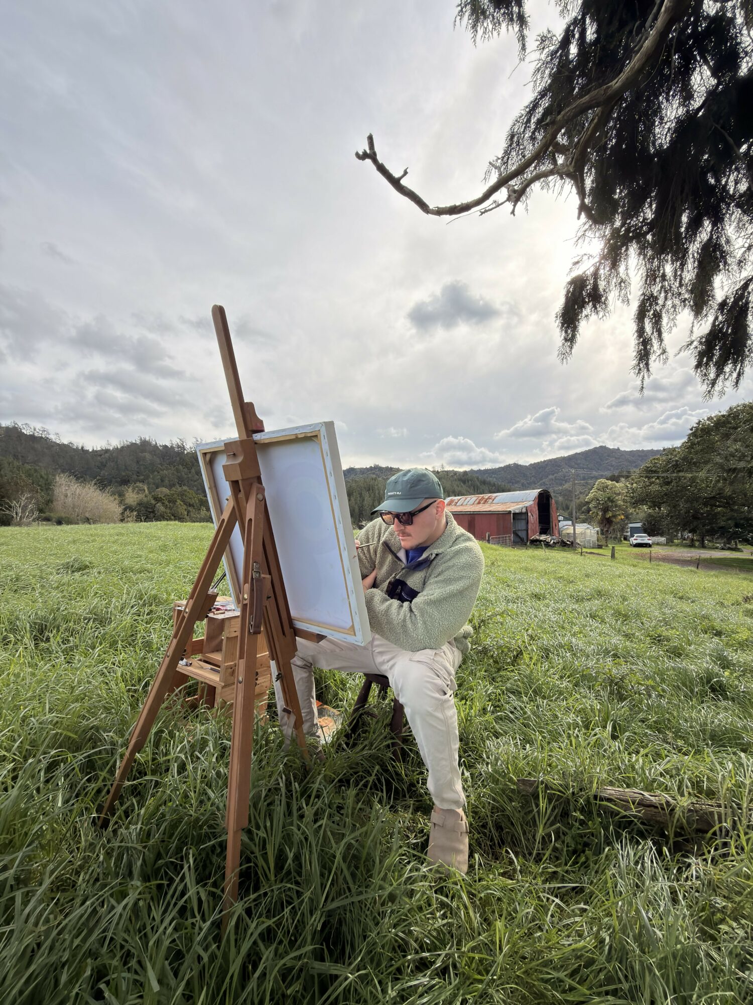 Person painting outdoors on an easel in a grassy field with trees and a barn in the background.