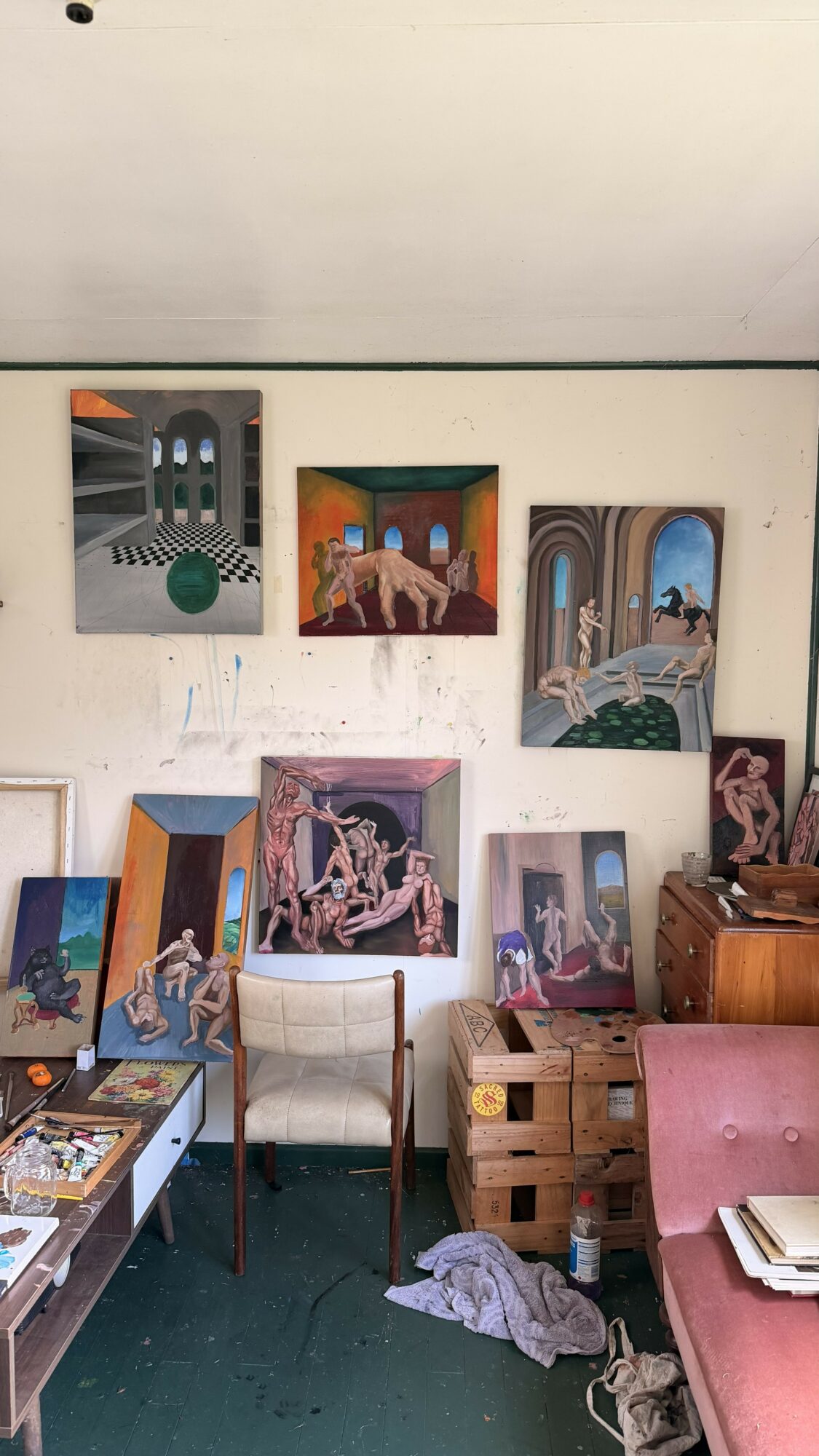 Wall with seven colorful paintings, a chair, a wooden crate, and a pink sofa with books and clothes nearby.