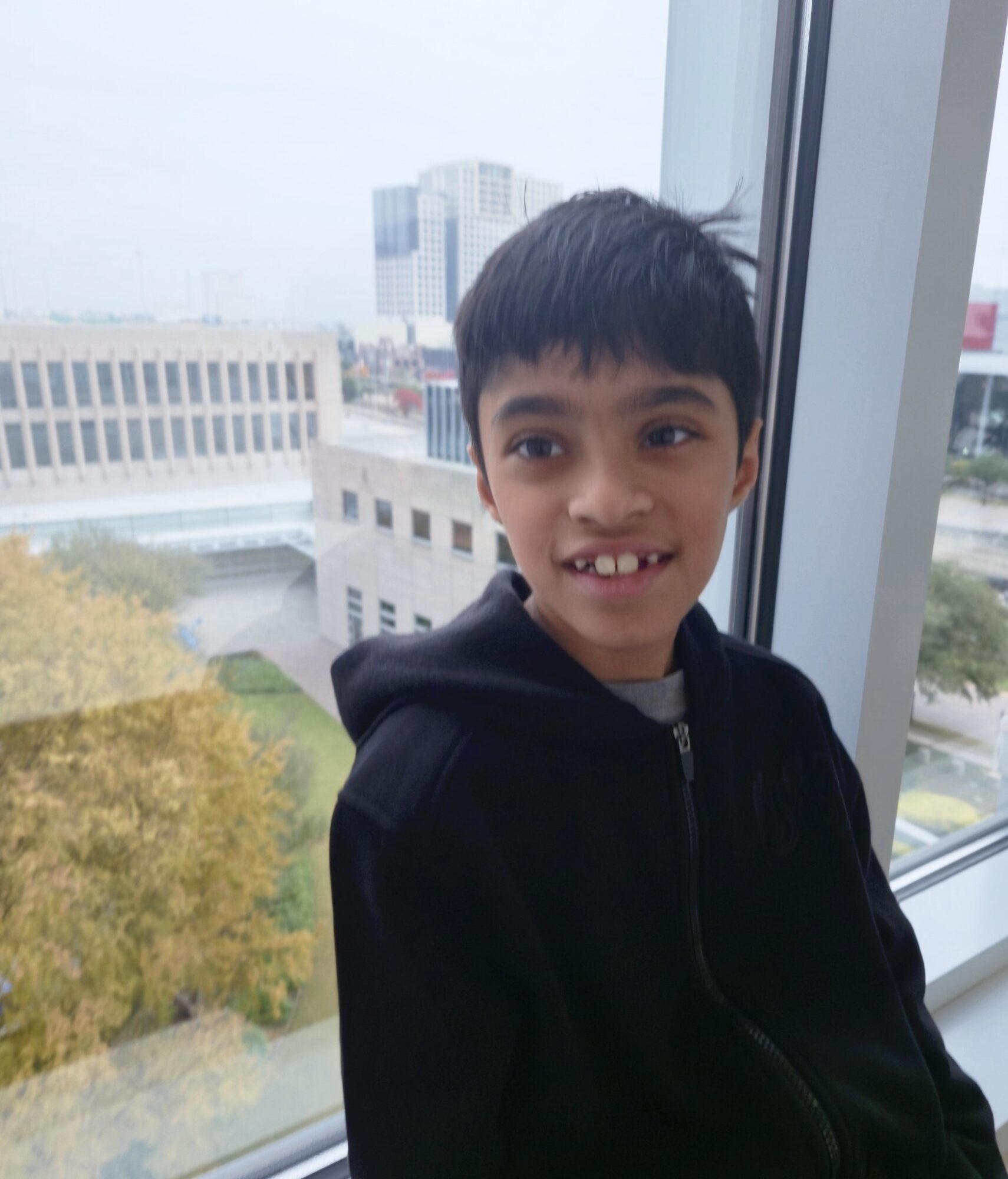 Young boy with dark hair smiling, sitting by a window with city buildings outside.