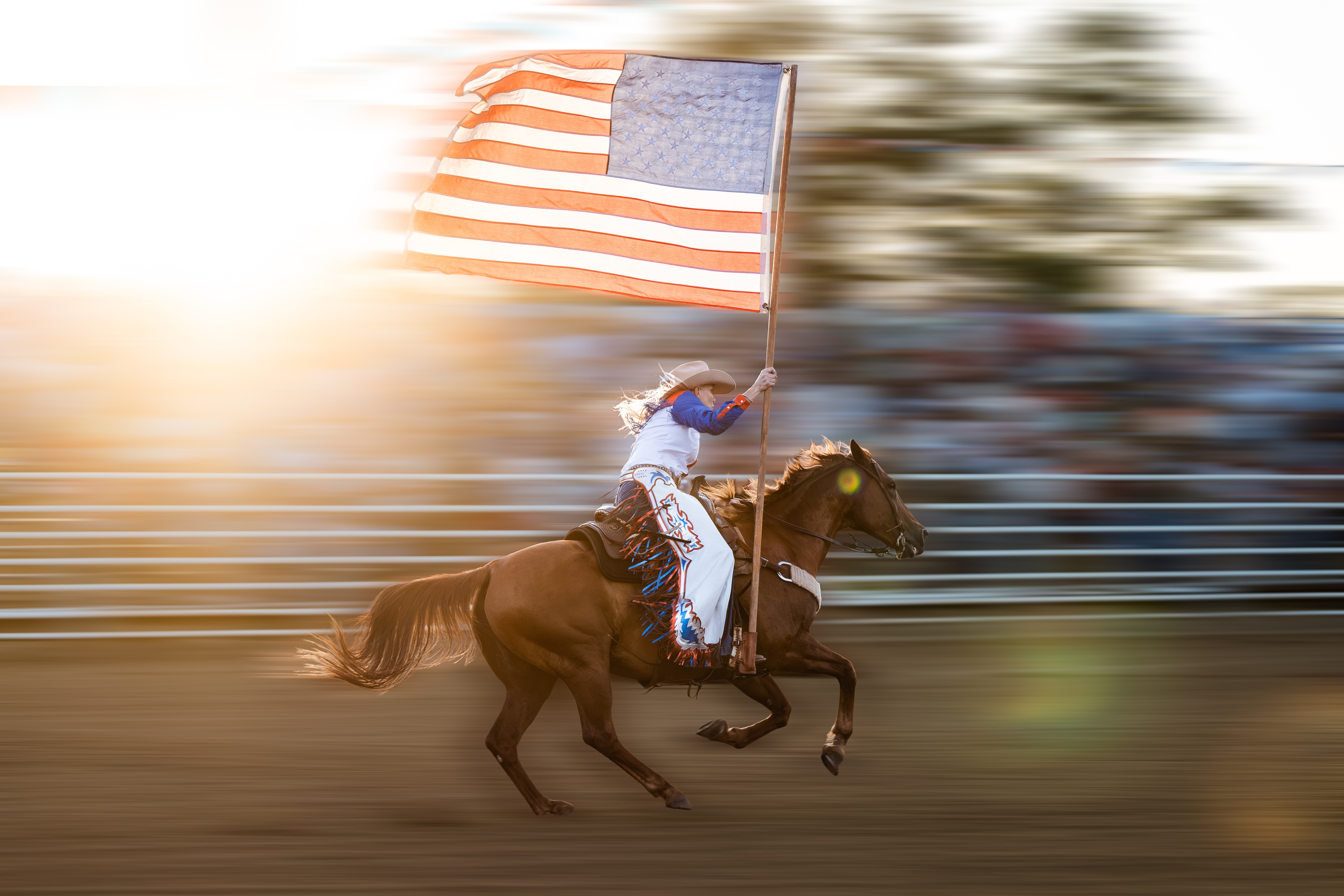 Person riding a horse while holding an American flag, with motion blur background, sunlight, and a fence.