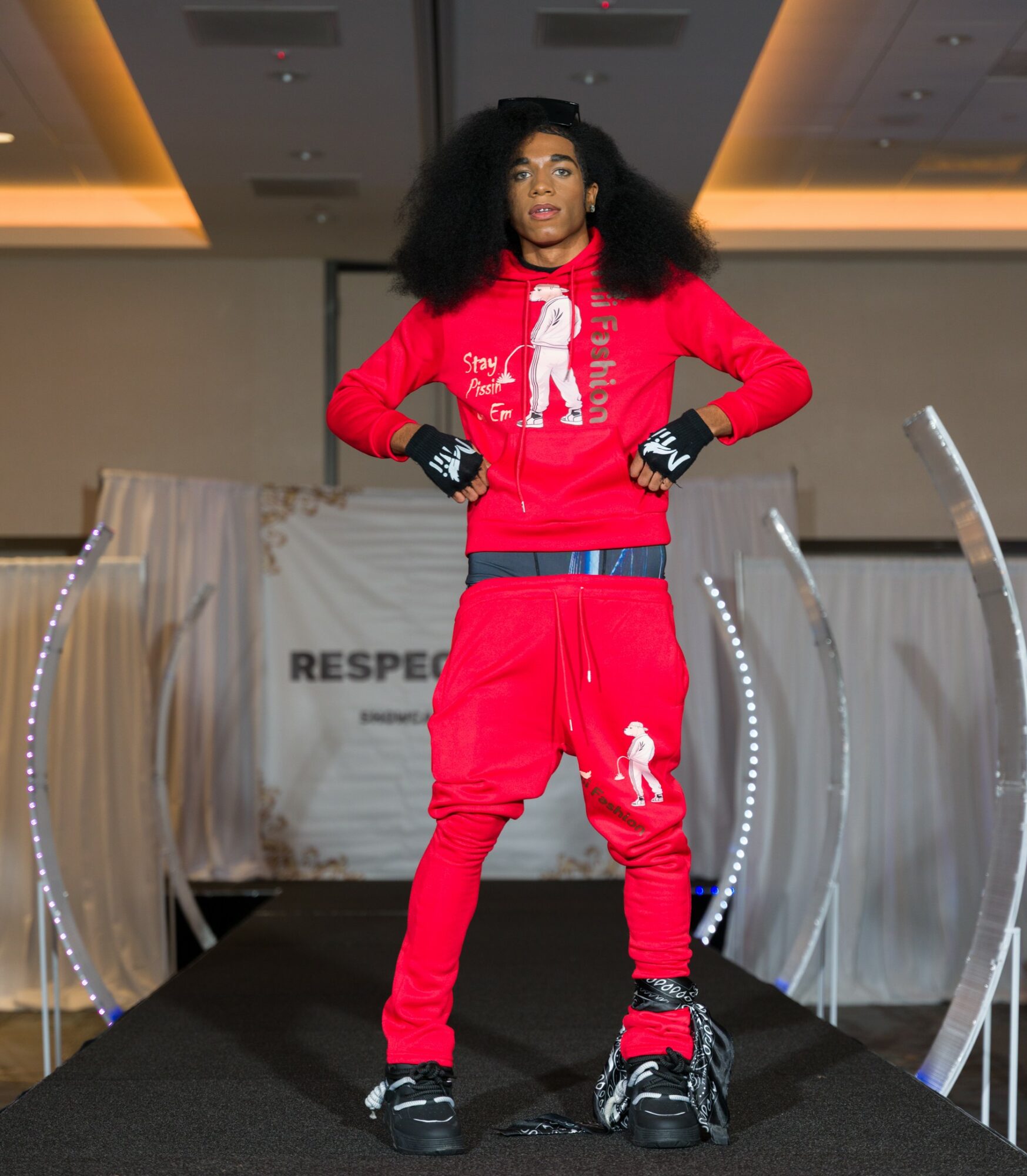 Woman in red athletic outfit with gloves and backpack on runway, with stage and lighting in background.