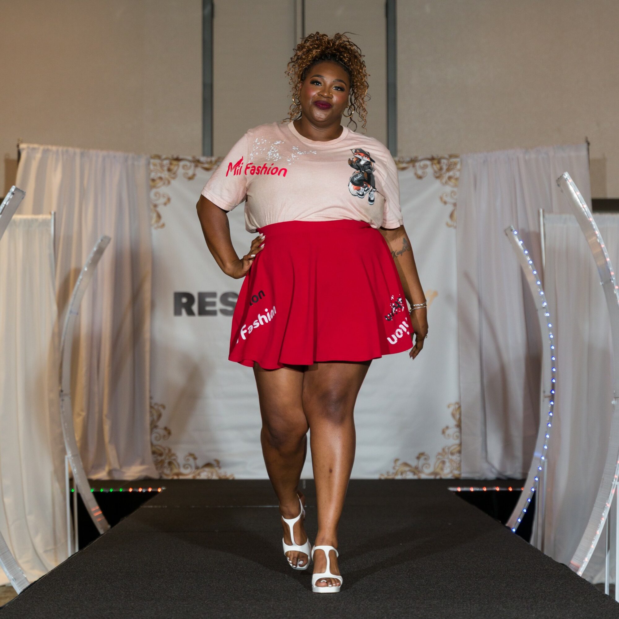 Woman walking on runway wearing a white top and red skirt, smiling, with a backdrop and runway lights.