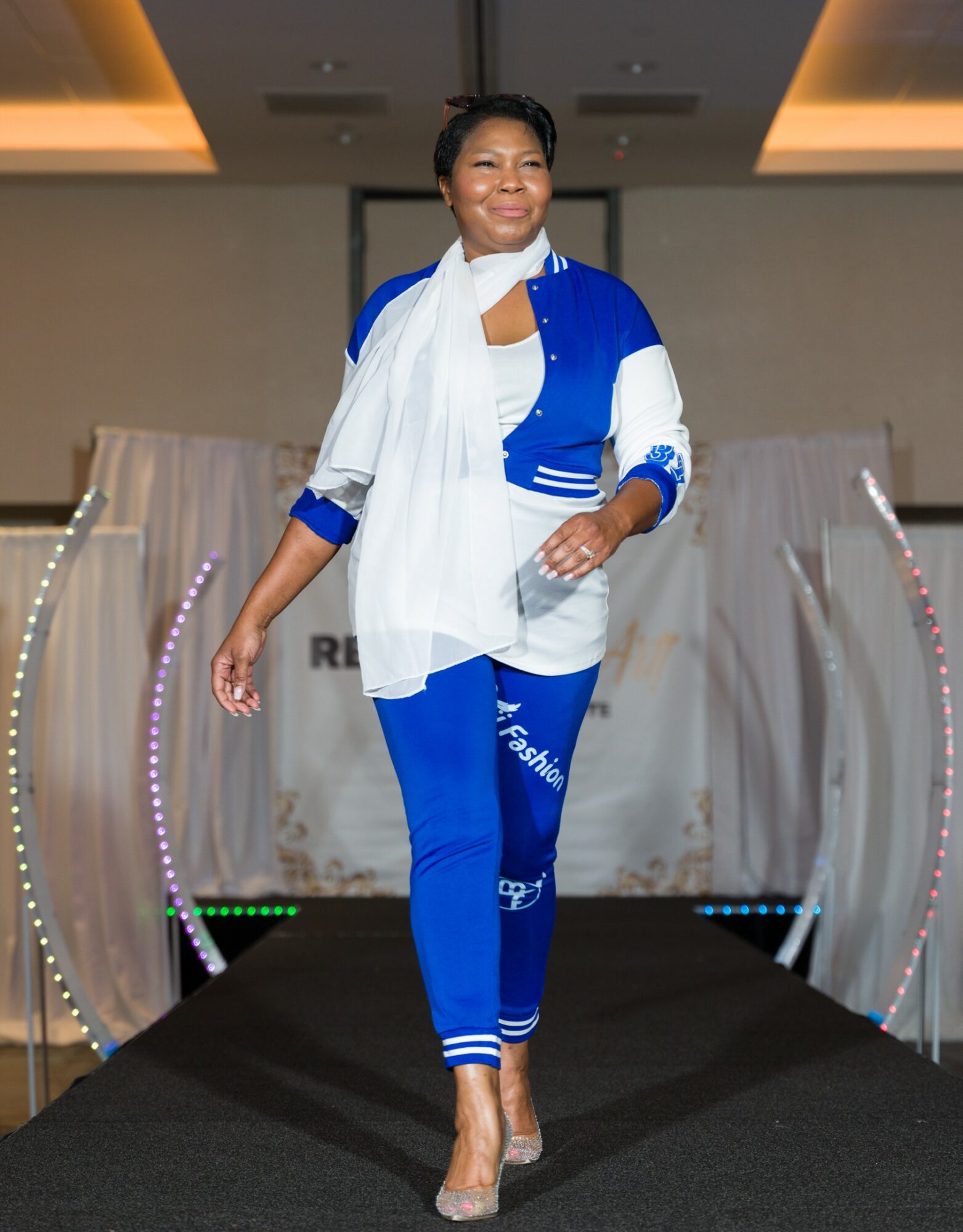 Woman walking on runway wearing blue and white outfit, smiling, with a stage background and decorative arches.