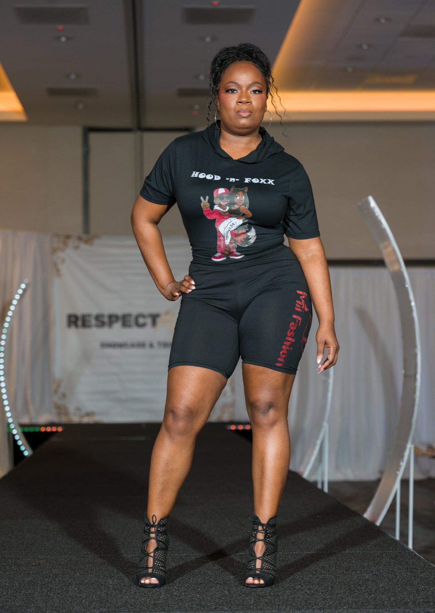 Woman standing confidently on runway, wearing black graphic t-shirt and shorts, with hands on hips, in an indoor event space.