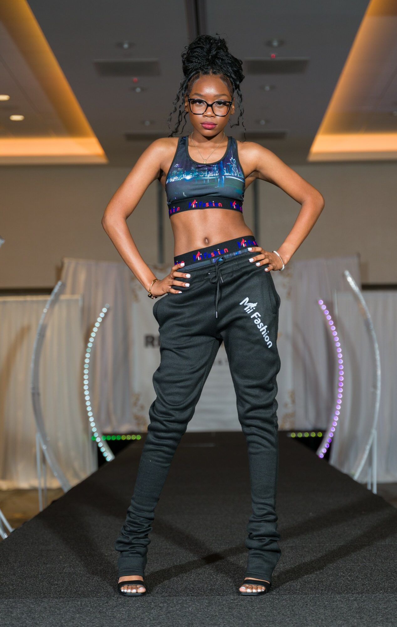 Young woman with glasses and braided hair stands confidently on runway, hands on hips, in a blue sports bra and black pants.