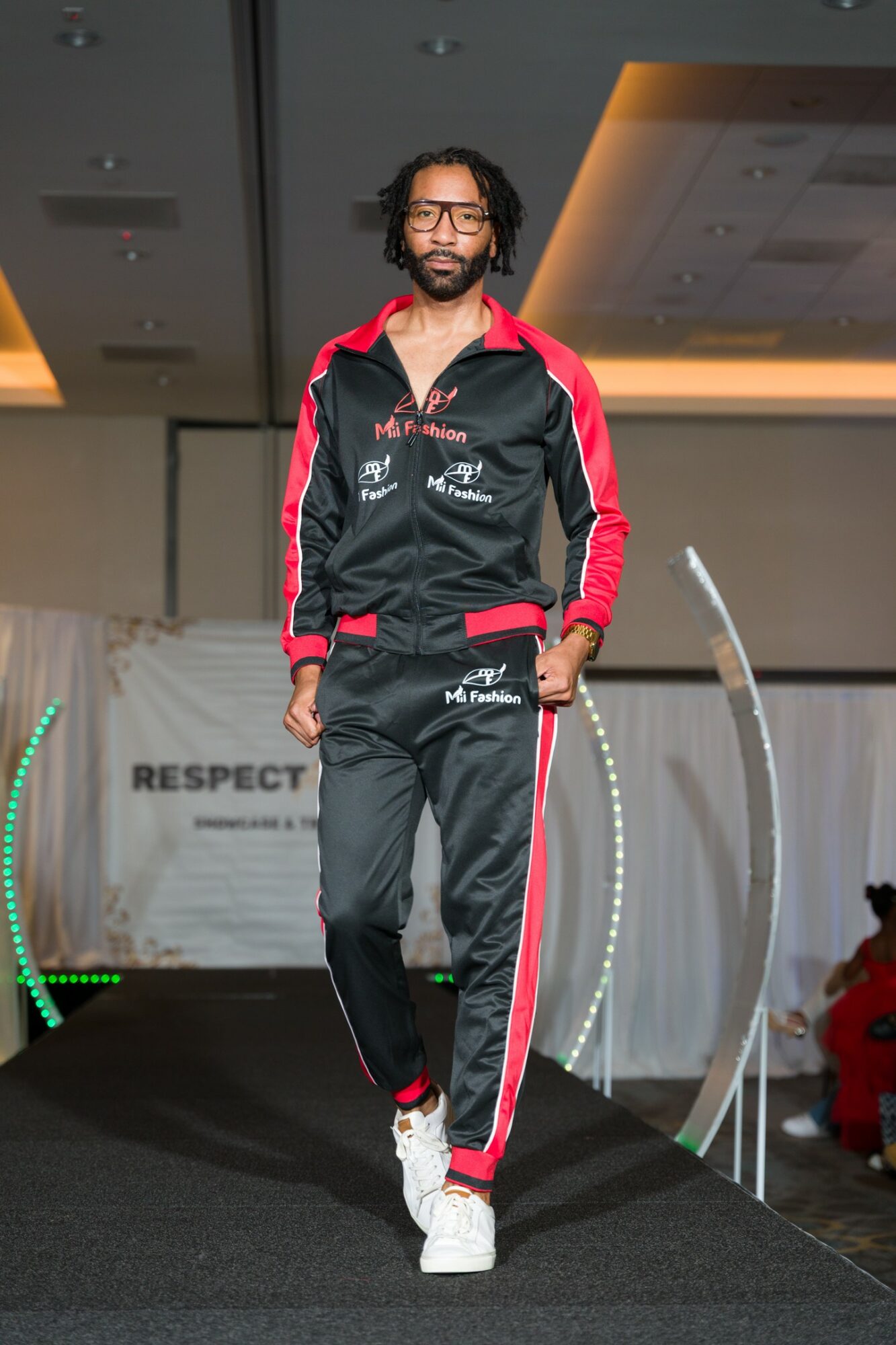 Man with glasses and curly hair walks on stage wearing a black and red tracksuit with white sneakers.
