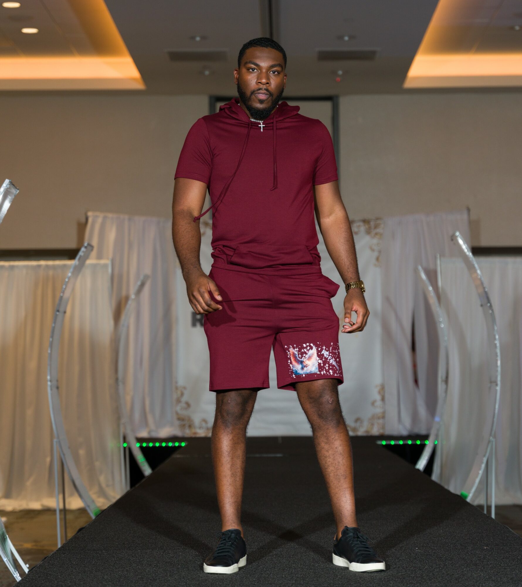 Man walking on runway in maroon hoodie and shorts with floral pattern, black sneakers, in indoor setting.