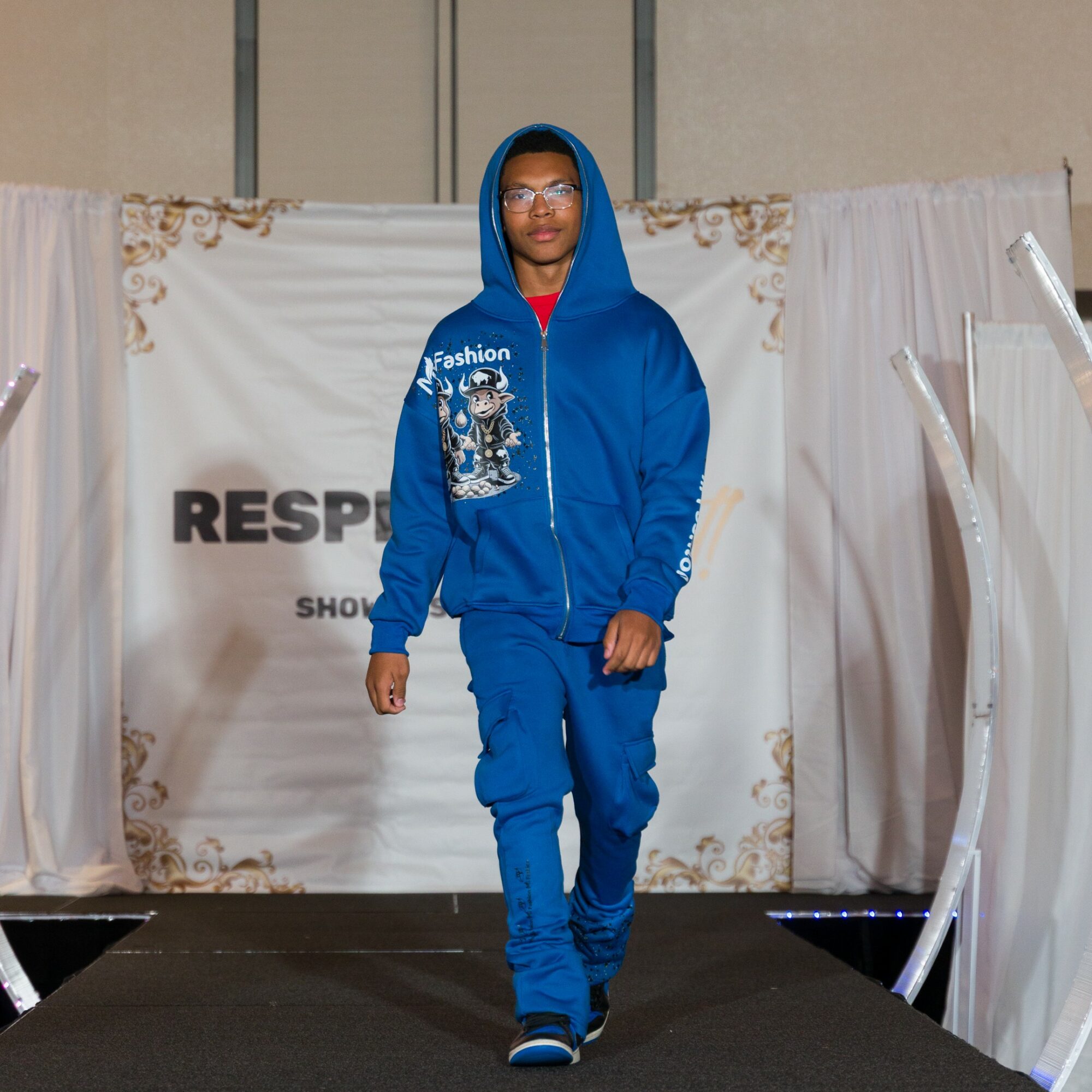 Person walking on runway wearing blue hoodie and pants, glasses, and glasses, with a backdrop reading RESPE, in an indoor setting.