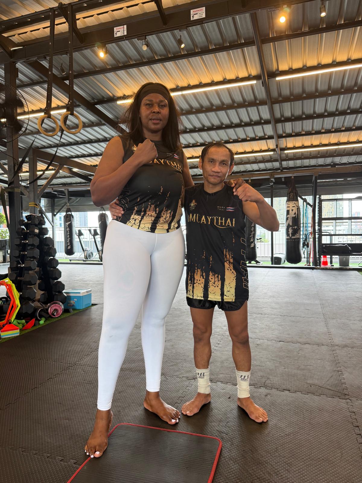 Two women standing in a gym, one with arm around the other, both smiling, with gym equipment in background.