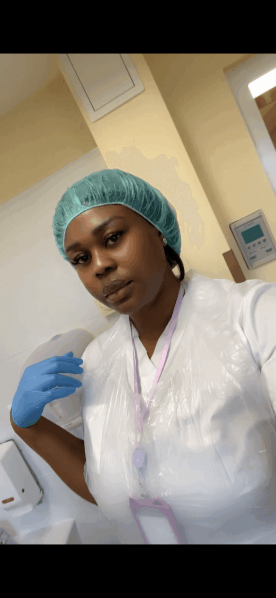 Healthcare worker wearing a green cap, white gown, and blue gloves in a clinical setting.