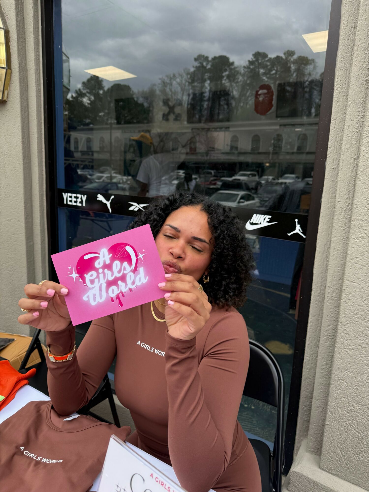 Young woman with curly hair holding a pink sign that says 'Fierce and Unapologetic' inside a store, with a window showing parking lot outside.