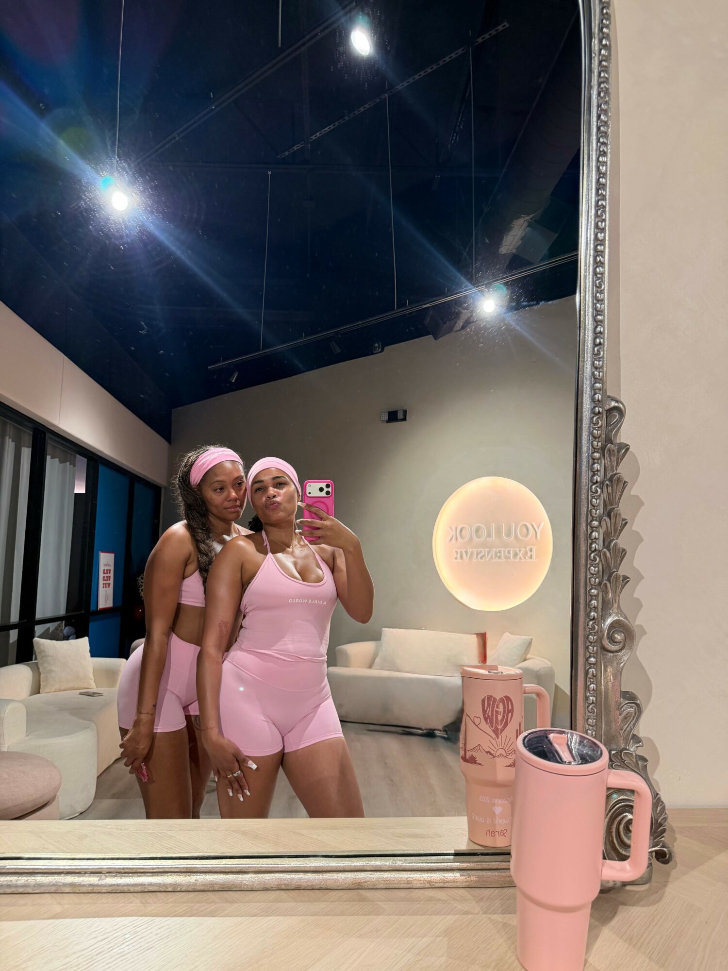 Two women in pink outfits taking a mirror selfie in a modern room with a large mirror, white couches, and decorative elements.