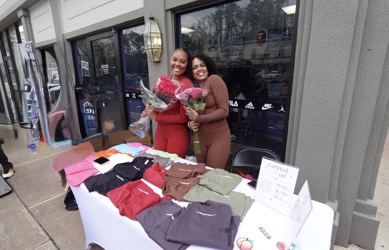 Two women smiling and hugging, one holding a bouquet of flowers, standing behind a table with folded clothing items outside a store.