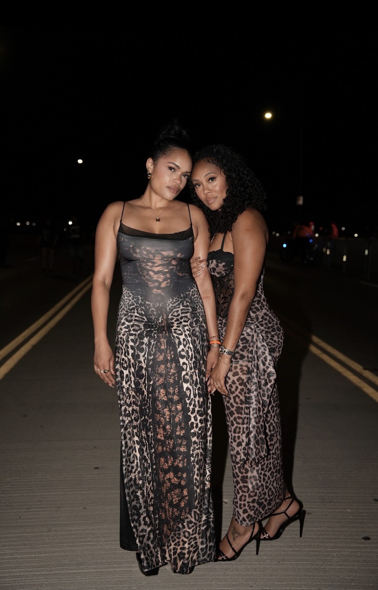 Two women in animal print dresses standing on a dark street at night, illuminated by streetlights, with one woman leaning on the other.