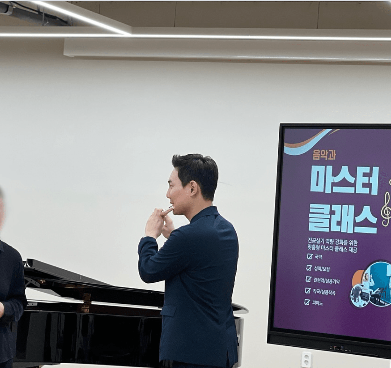 Man in dark suit standing near a piano and a presentation screen, in a room with white walls.