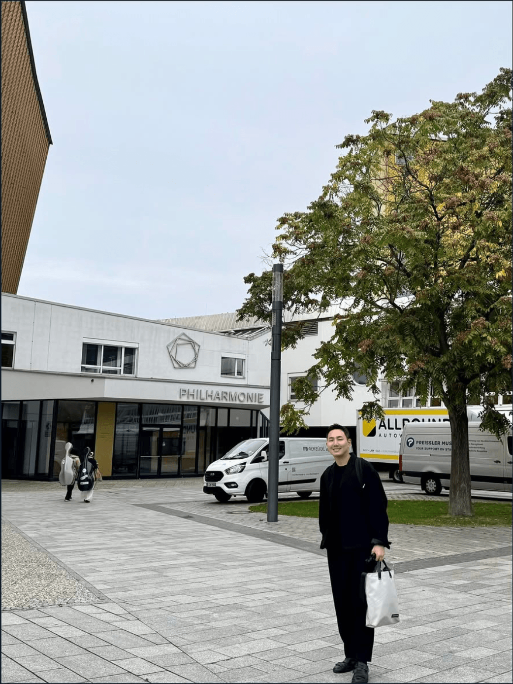 Person standing outdoors near a tree and building, with a white van and a few people in the background.