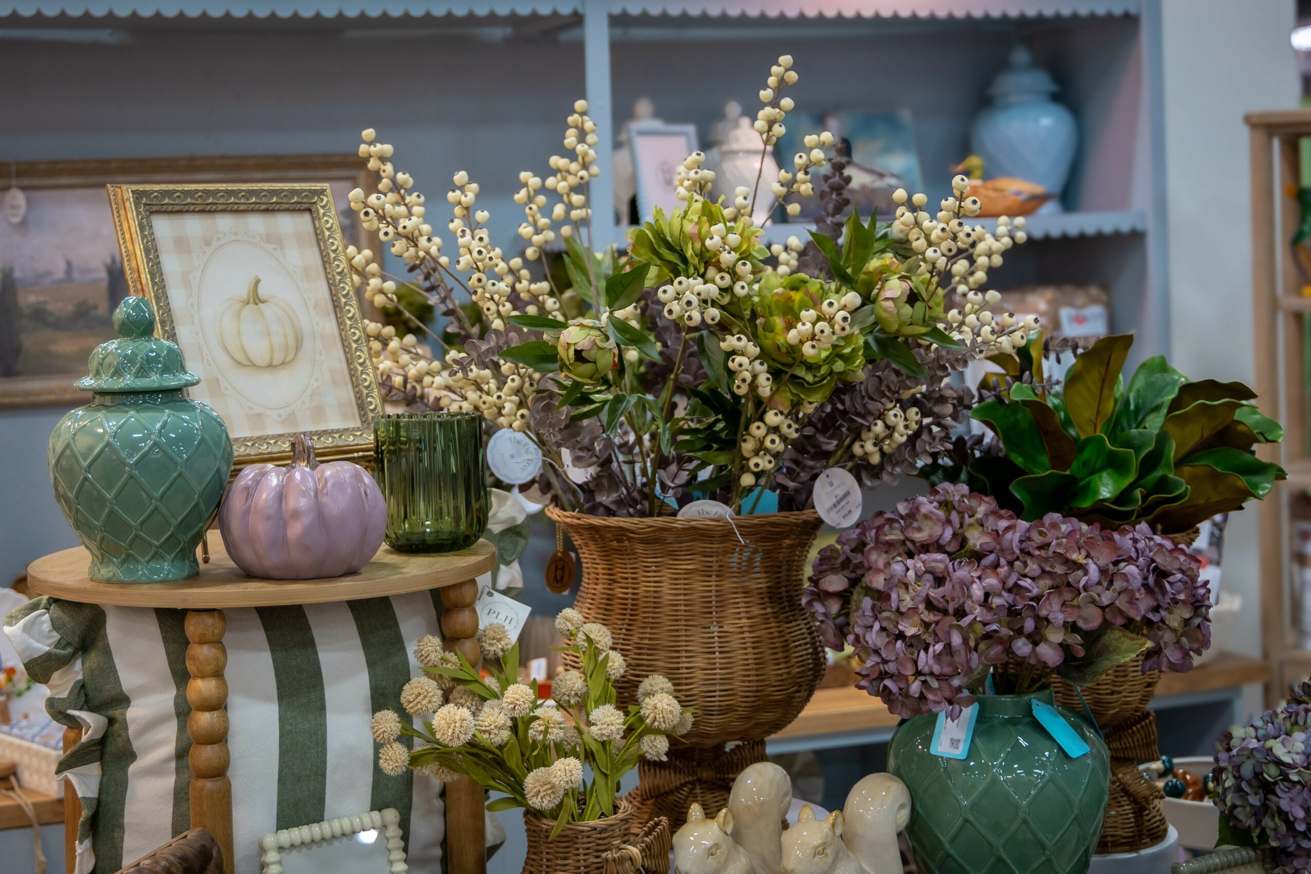 Vase with white and purple flowers, surrounded by decorative items and a striped tablecloth, in a room with shelves.