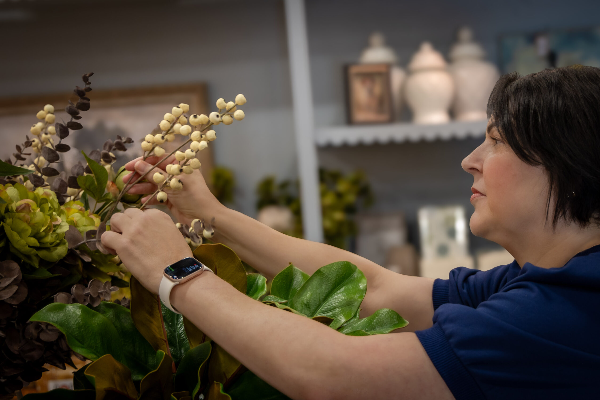 Woman arranging flowers in a room with shelves and decorative items in the background.