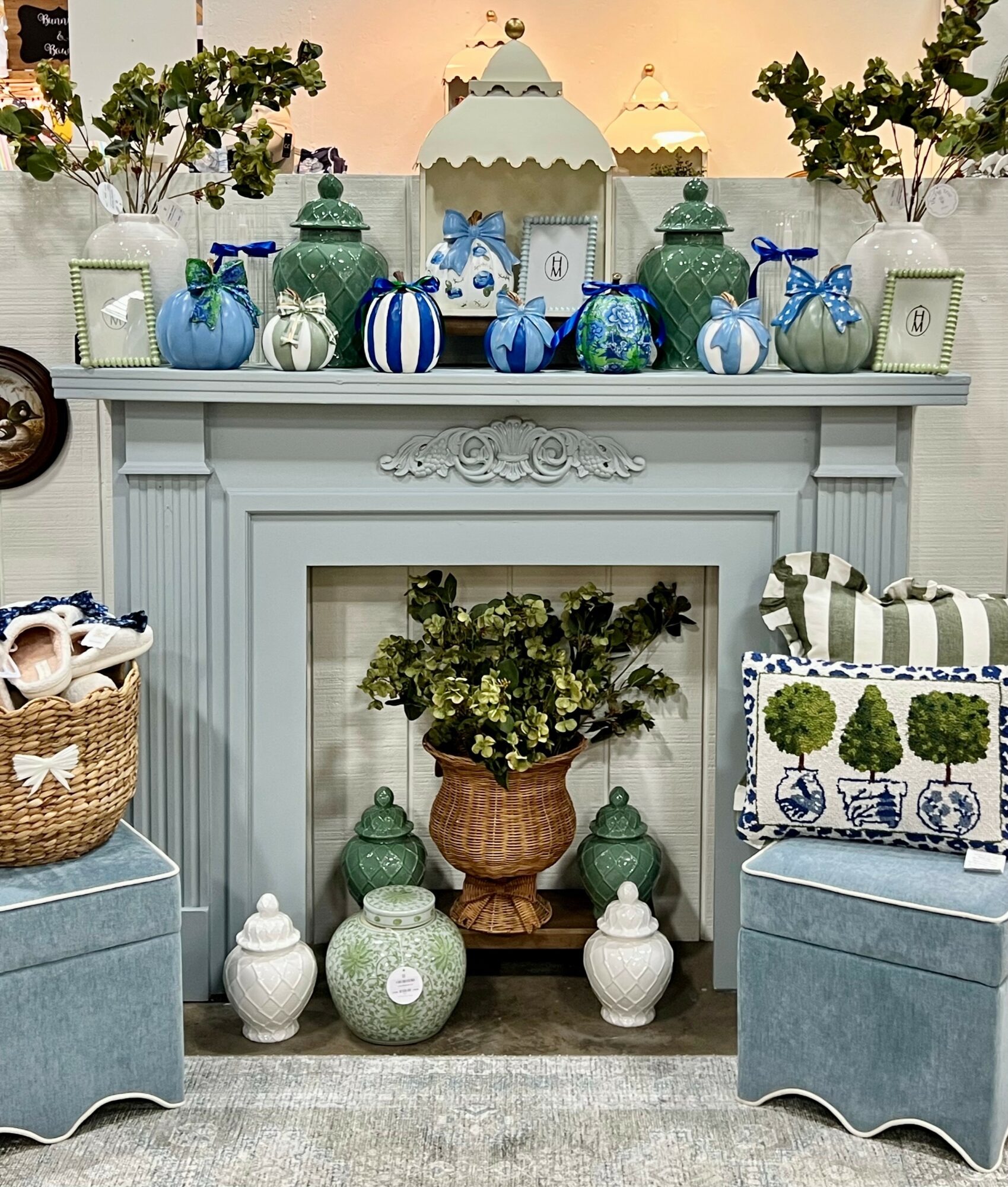 Decorative fireplace mantel with blue and green vases, plants, and ornaments, flanked by blue storage chests, with a floral arrangement inside the fireplace opening.