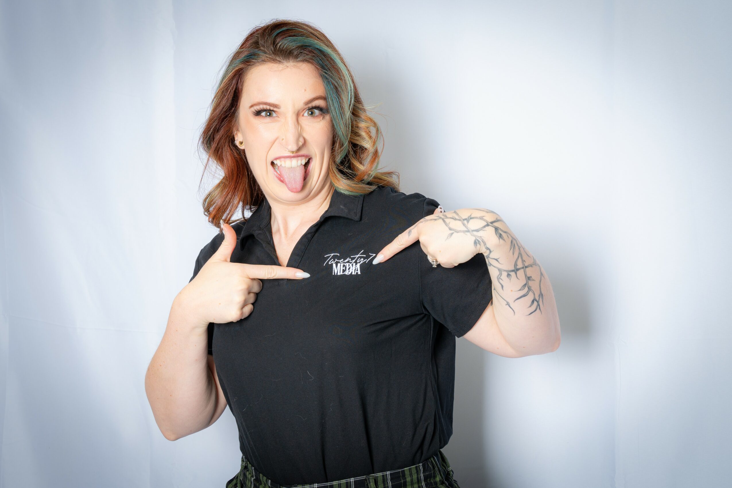Woman with colorful hair pointing to her black shirt, sticking out her tongue, and showing a tattoo on her arm.