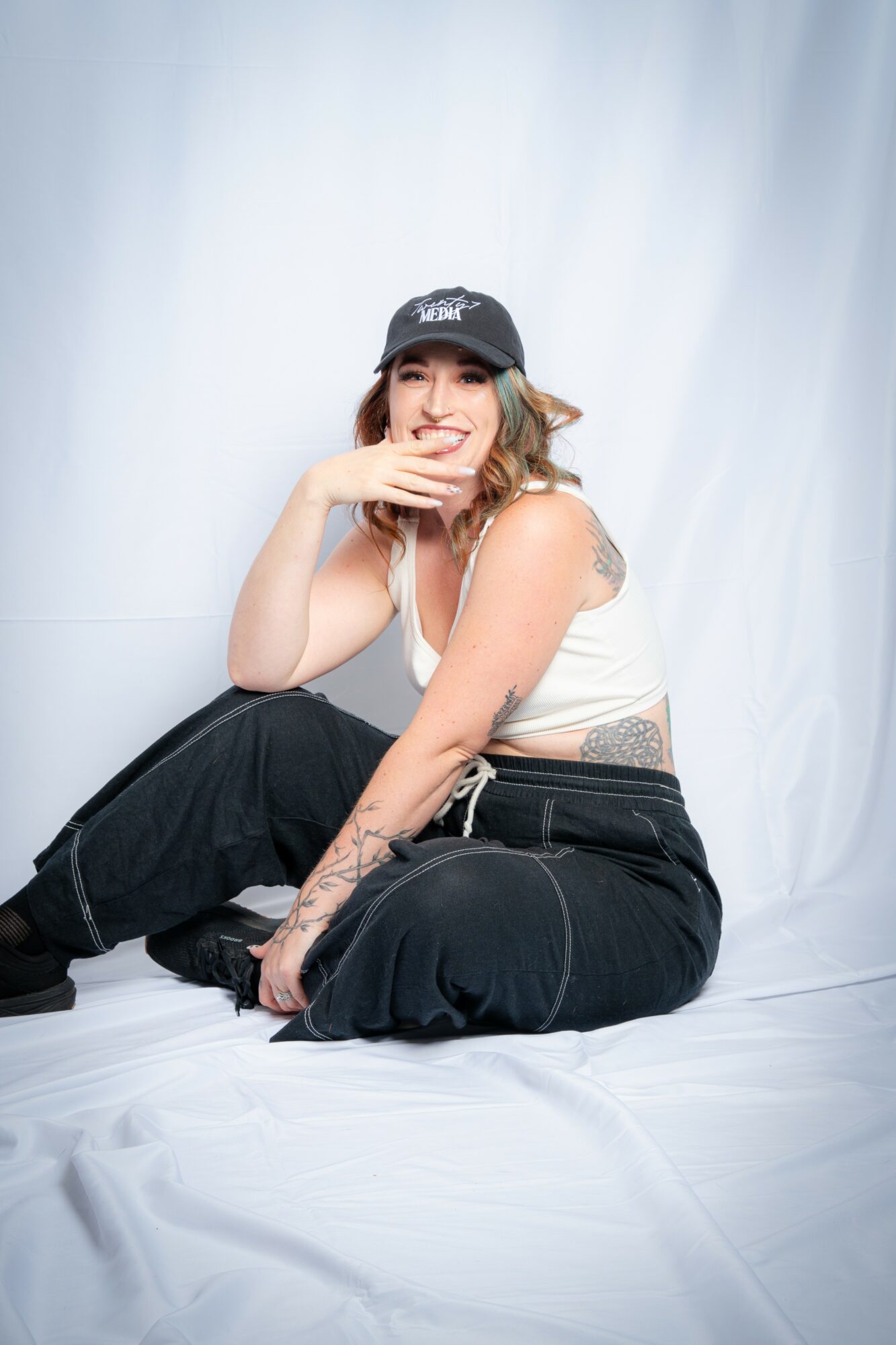 Young woman sitting on the floor, smiling, wearing a black cap, white top, and black pants, with a white backdrop.