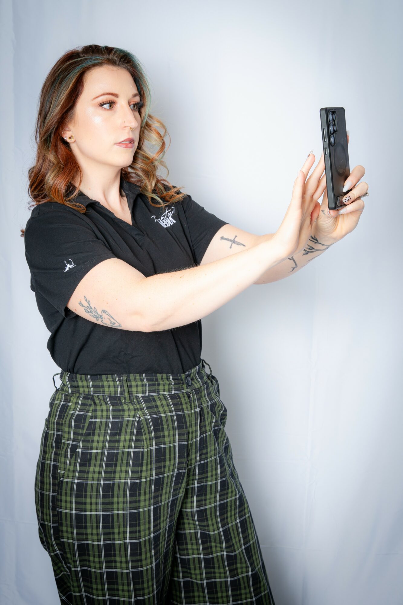 Woman with shoulder-length hair taking a selfie with a smartphone, wearing a black shirt and plaid pants, standing against a plain background.