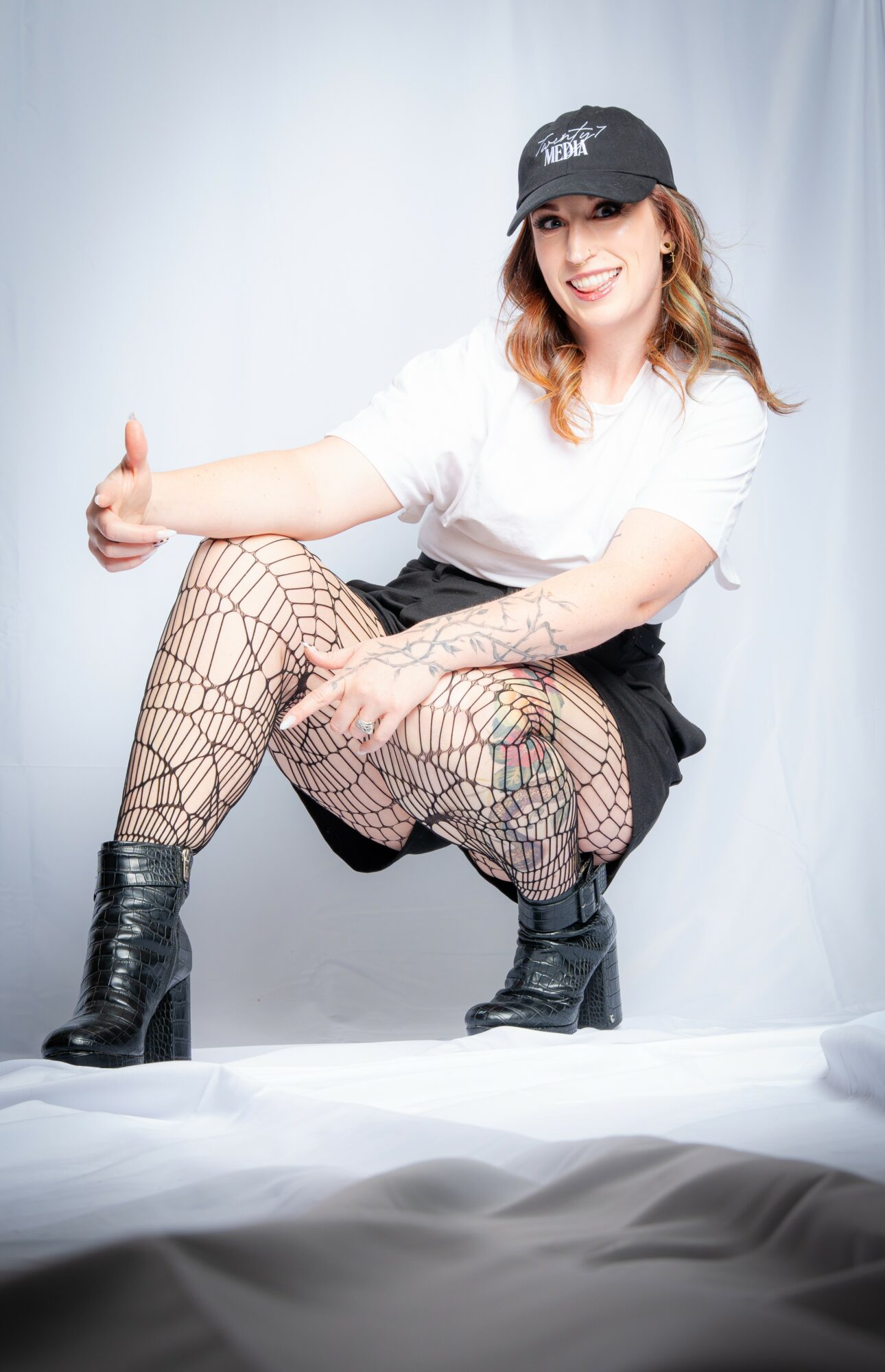 Young woman crouching, smiling, wearing a cap, white t-shirt, patterned fishnet tights, black skirt, and boots, giving a thumbs-up.