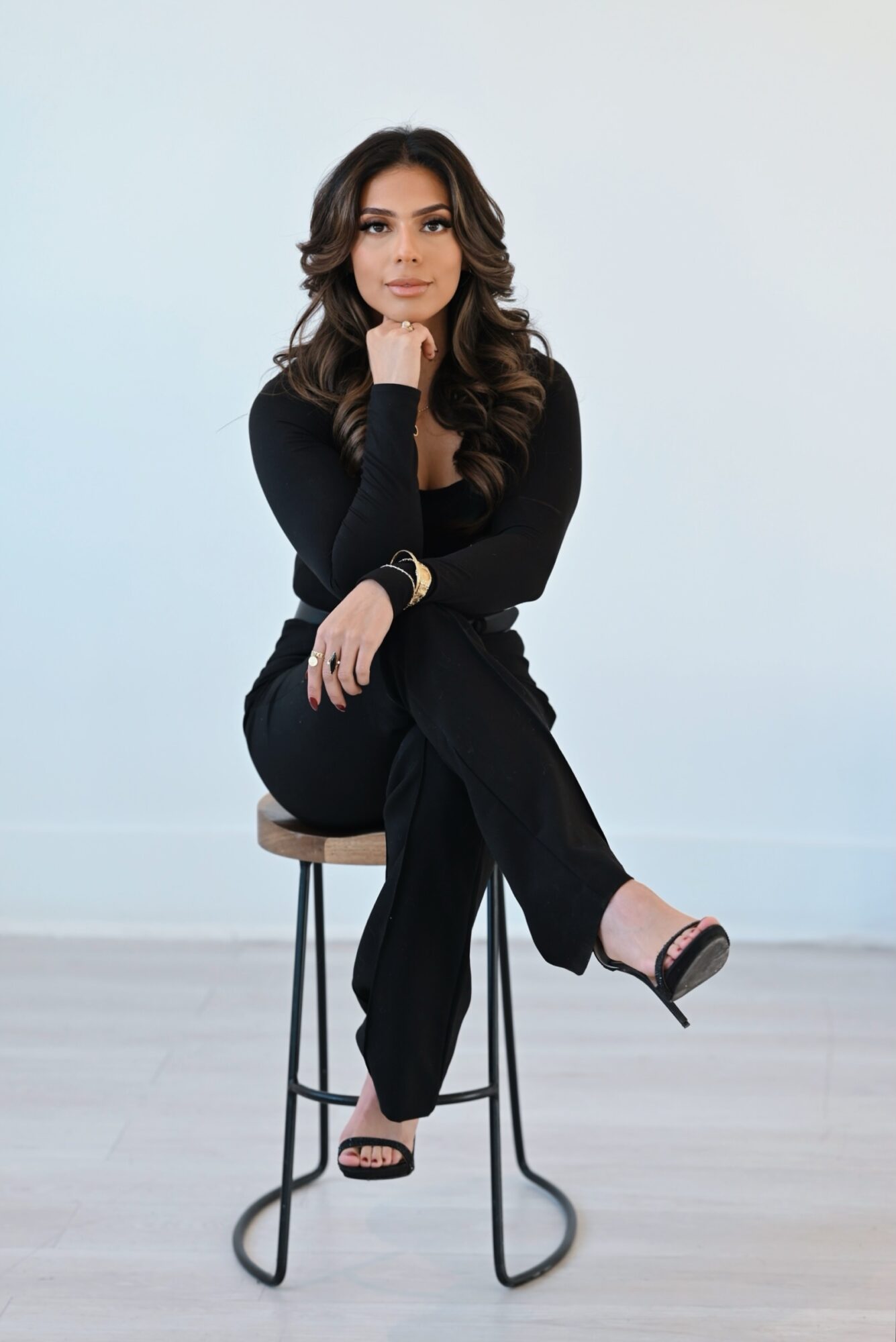 Woman with long dark hair sitting on a stool, wearing black outfit, against a light background.