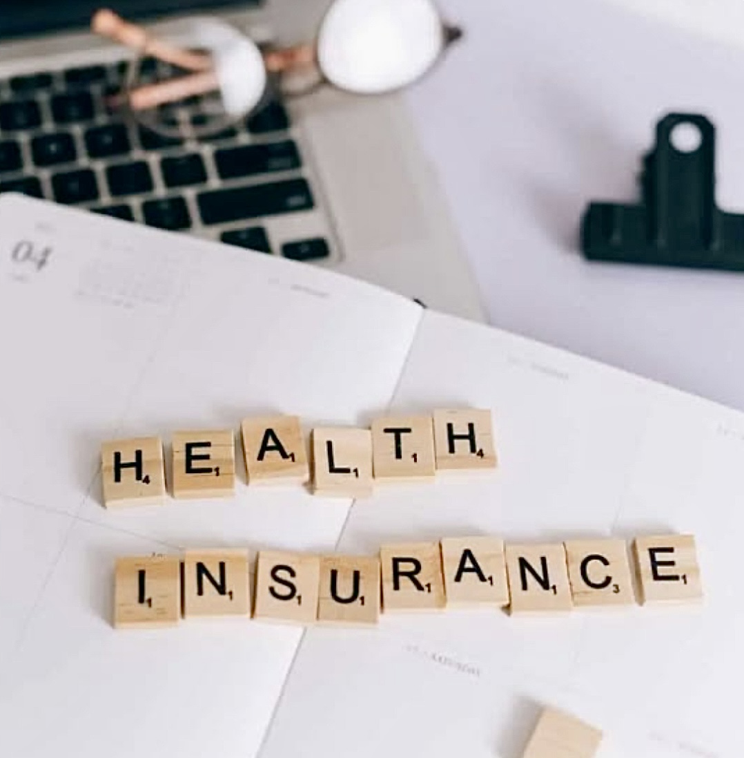 Scrabble tiles spell 'HEALTH' and 'INSURANCE' on a desk with a laptop, papers, and office supplies nearby.