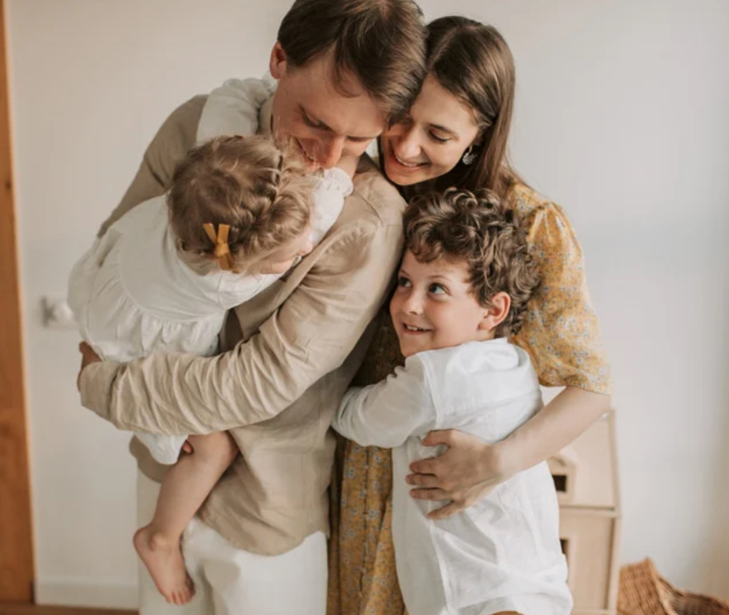 Family hugging indoors, two adults and two children smiling and embracing each other.