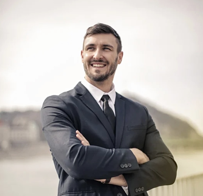 Man in a suit smiling outdoors with arms crossed, background of a river and hills