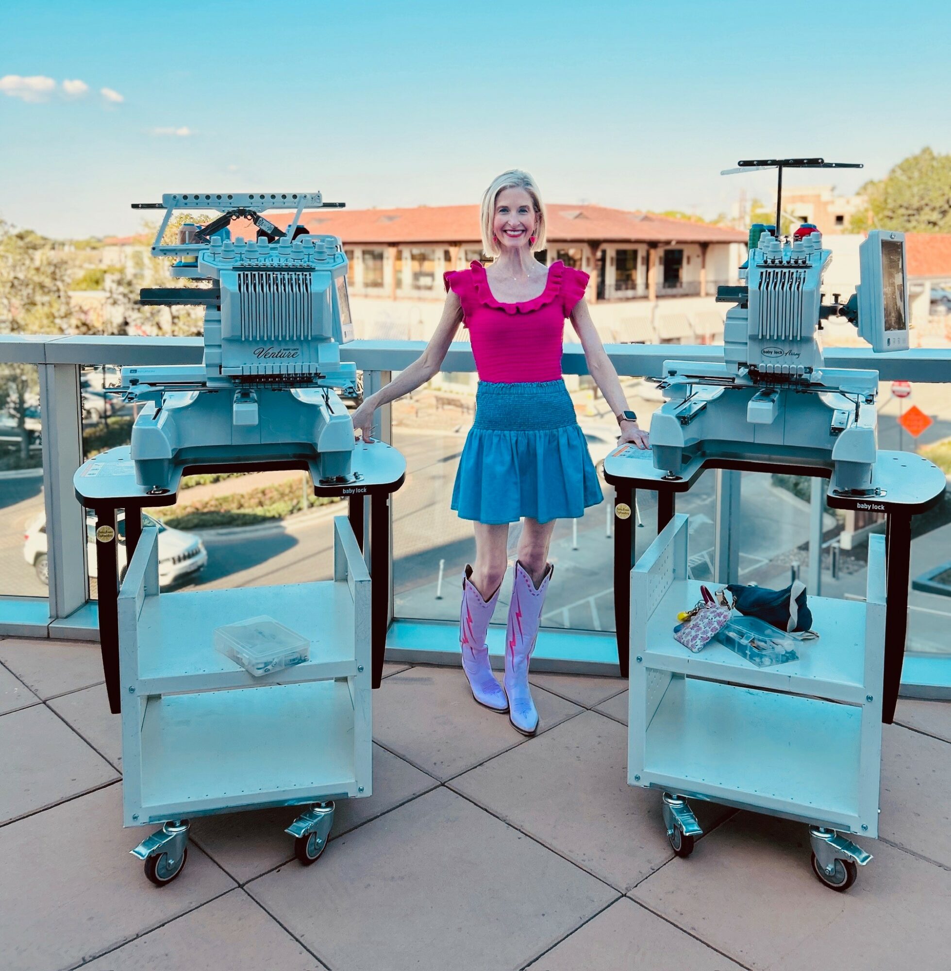 Woman standing between two large machines on a balcony, with a cityscape in the background, smiling, wearing a pink top and blue skirt.