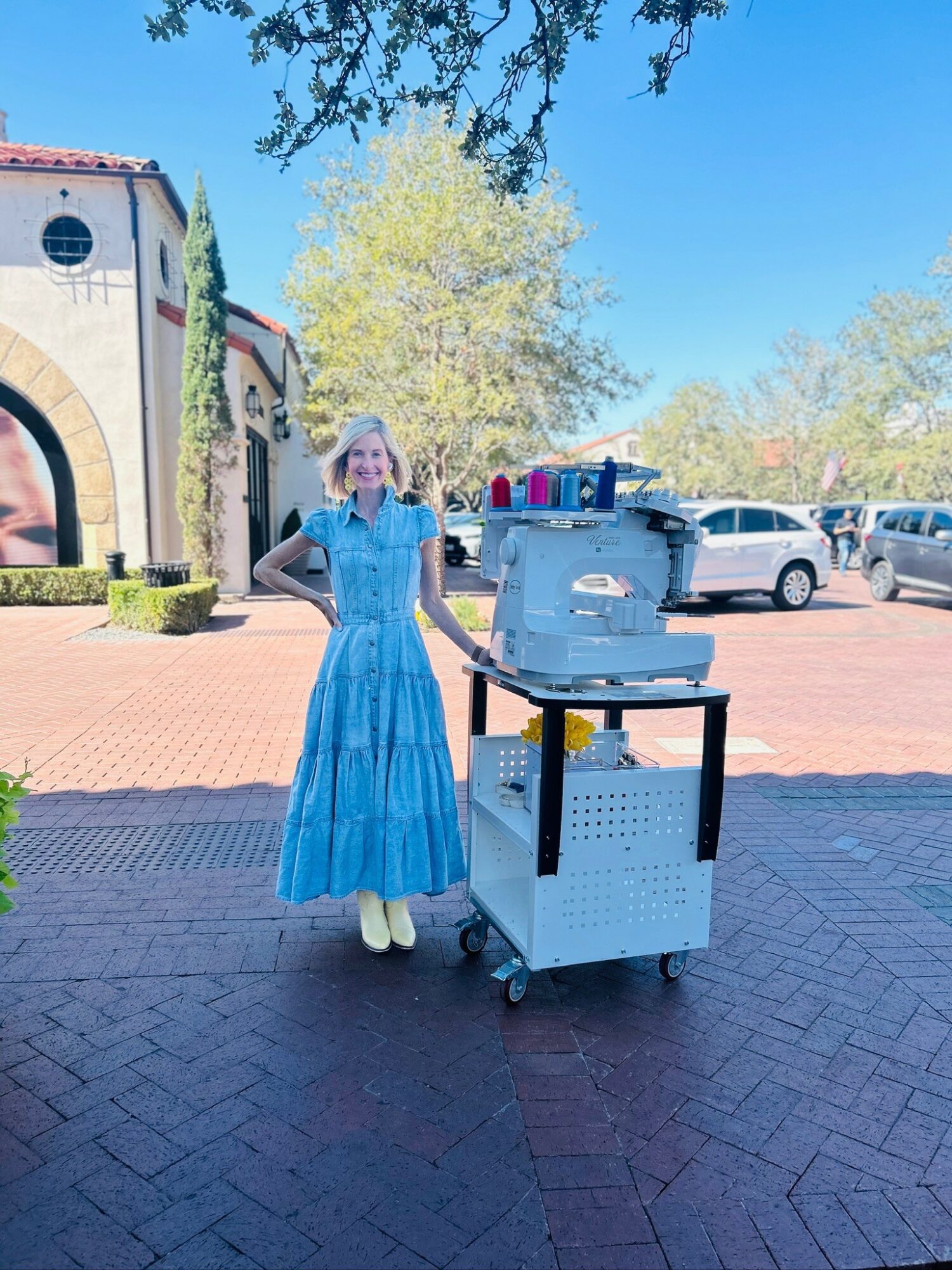 Woman in a blue dress standing outdoors next to a cart with beverage dispensers, trees, and parked cars in background.