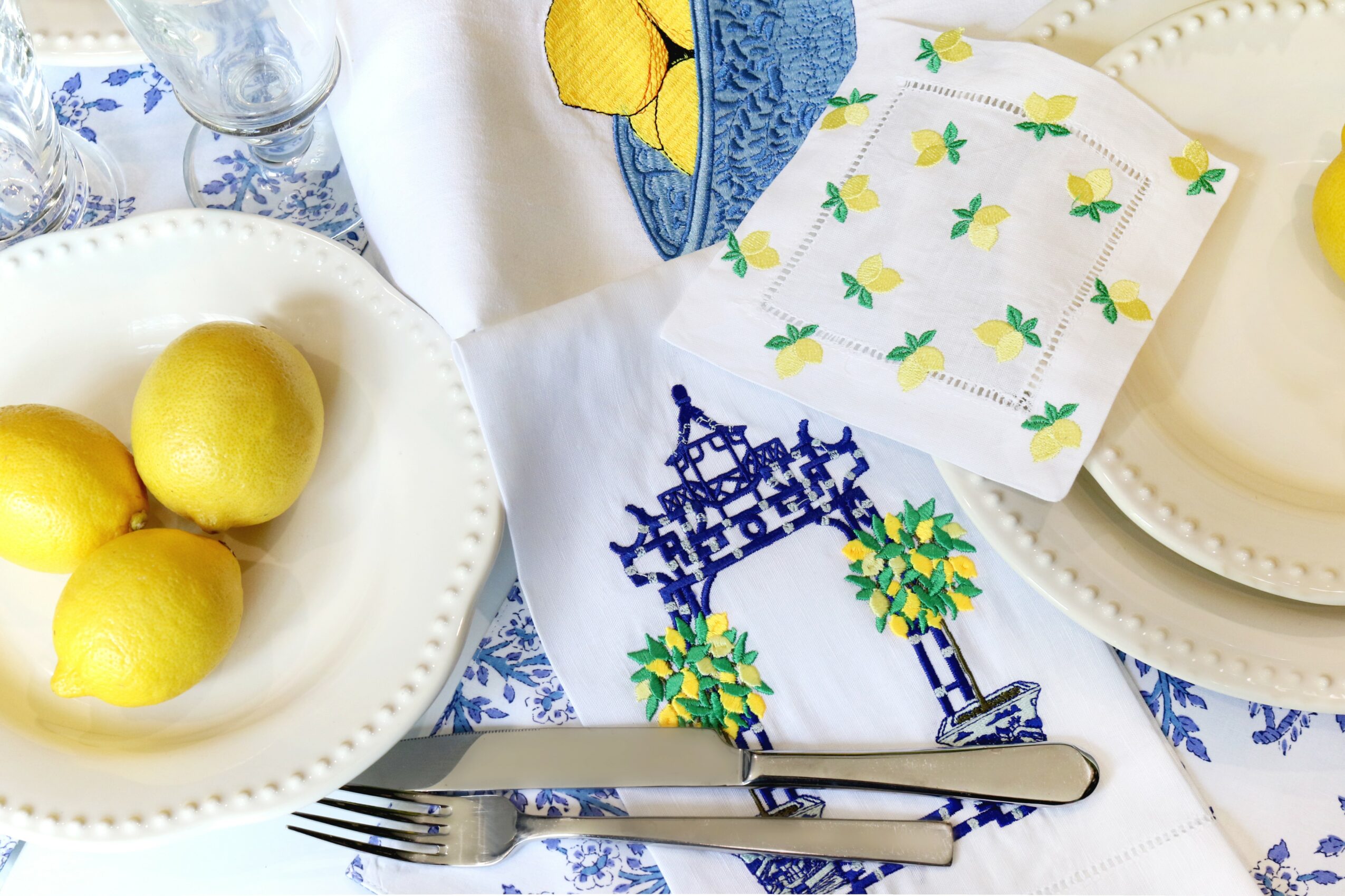 Lemon fruits on a white plate, surrounded by patterned cloths and napkins with blue and yellow designs, and a fork and knife.